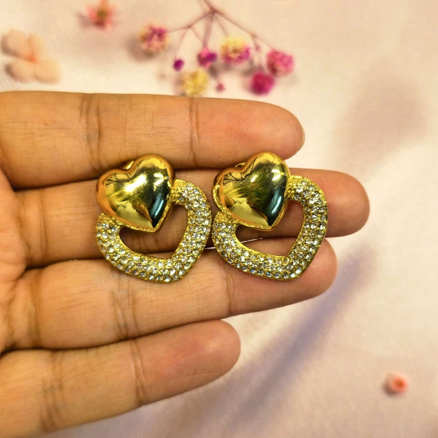 Gold heart-shaped earrings with gemstones held between fingers against a soft background.