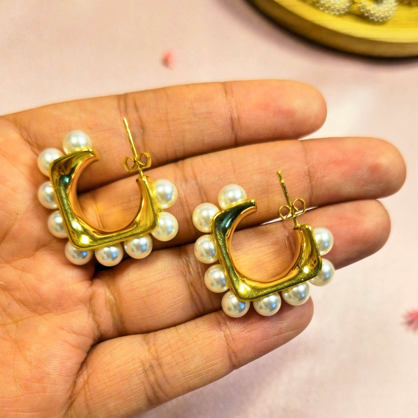 Gold hoop earrings with pearl accents held in a hand against a pink background