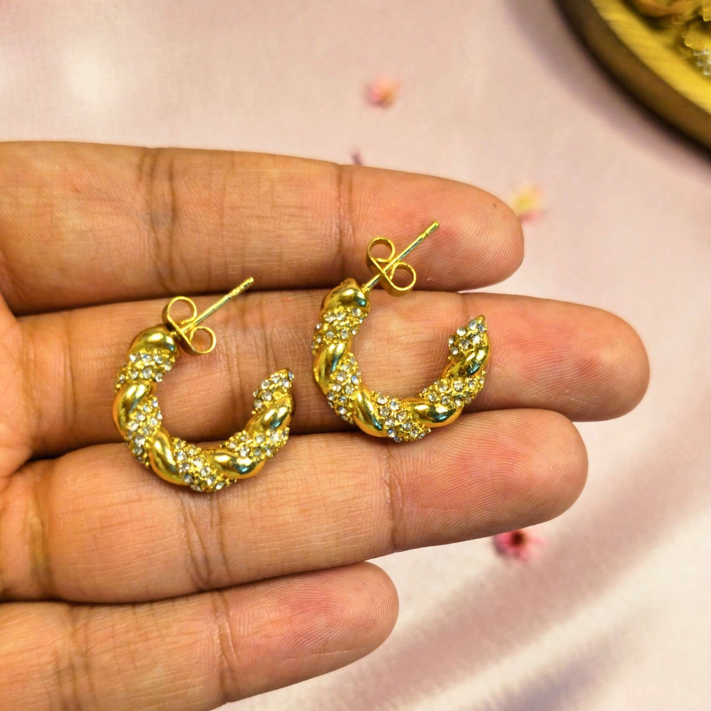 Gold diamond hoop earrings held between fingers against a pink background