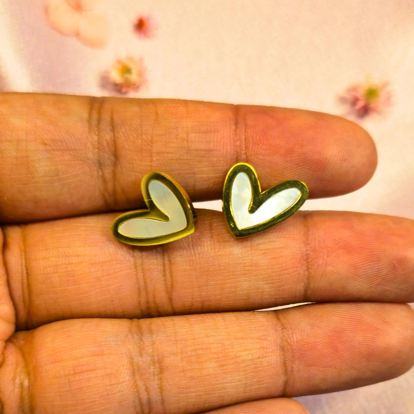 Gold heart-shaped white enamel centered earrings held between fingers with a blurred floral background