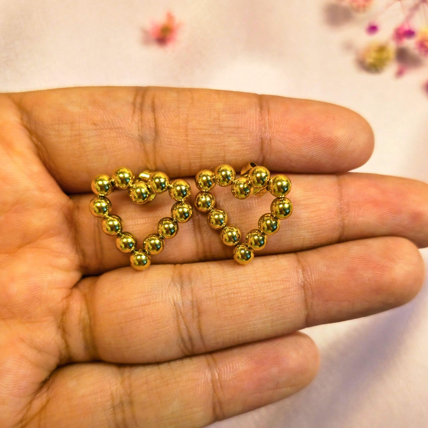 Gold heart-shaped beaded earrings held in a hand with a blurred floral background
