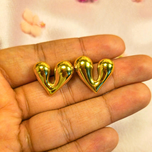 Gold heart-shaped earrings held between fingers against a neutral background