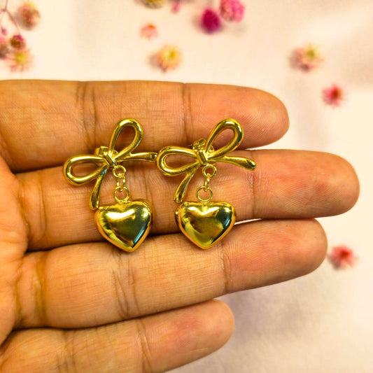 Gold bow and heart-shaped earrings held between fingers with a light background