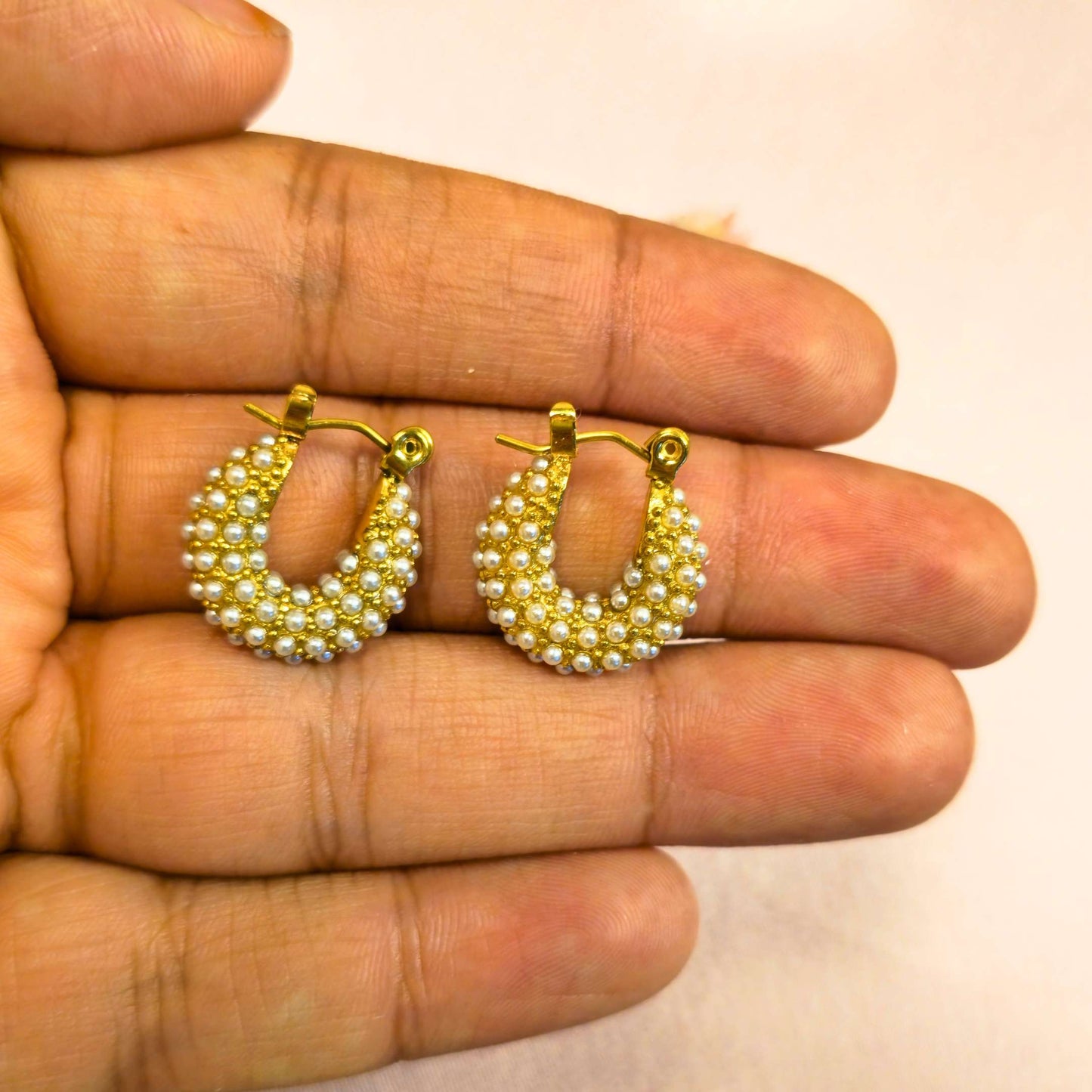 Gold hoop earrings with pearl-like beads held between fingers against a neutral background