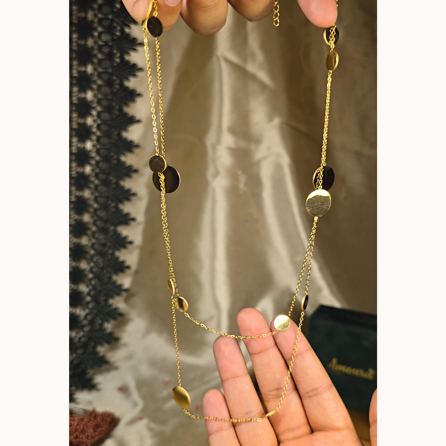 Gold necklace with black and gold circular pendants held by a hand against a neutral background
