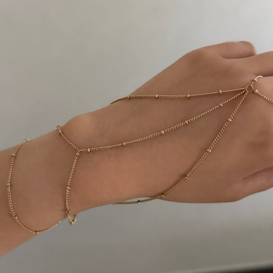 Hand wearing multiple gold ring chain bracelets on a neutral background