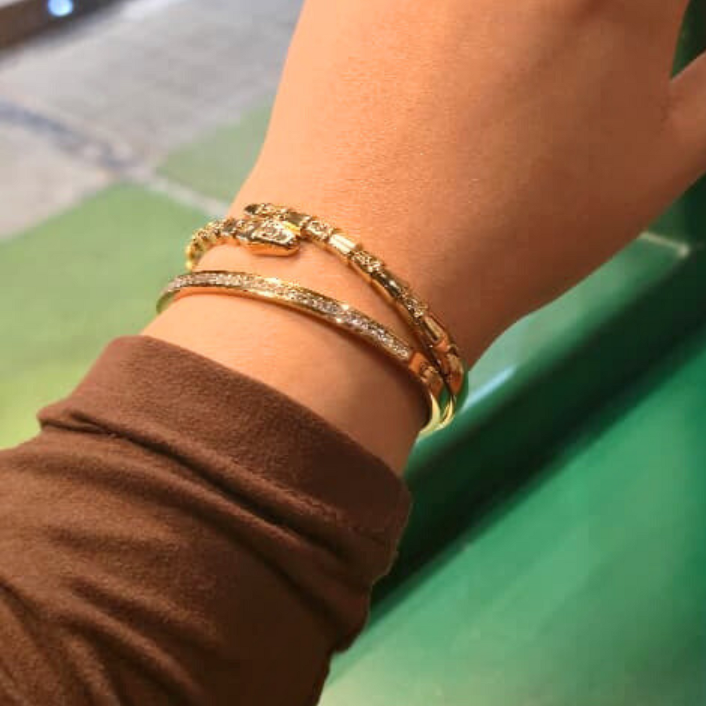 Close-up of a wrist wearing two gold bracelets with a blurred green background
