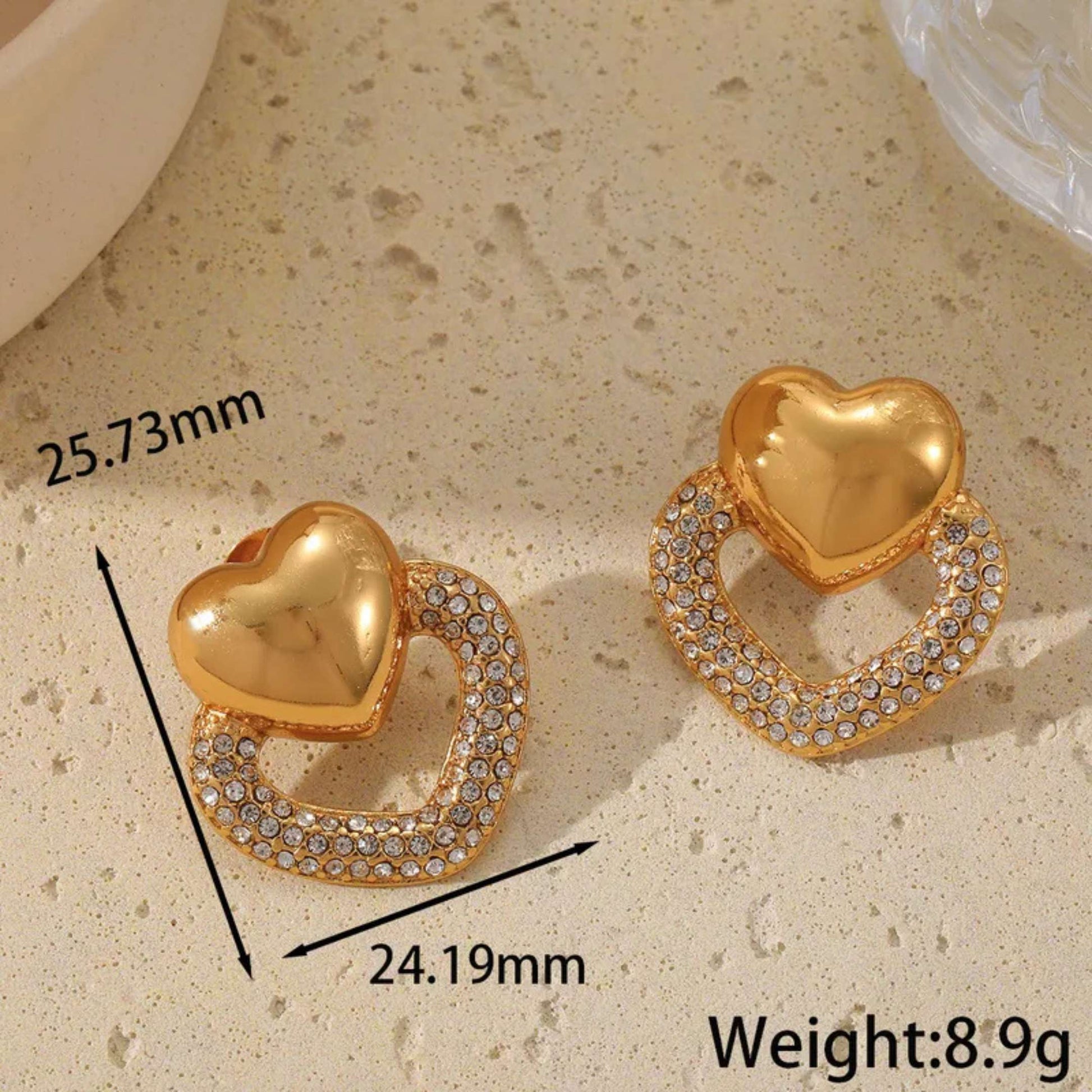 Gold heart-shaped earrings with measurements and weight on a beige surface
