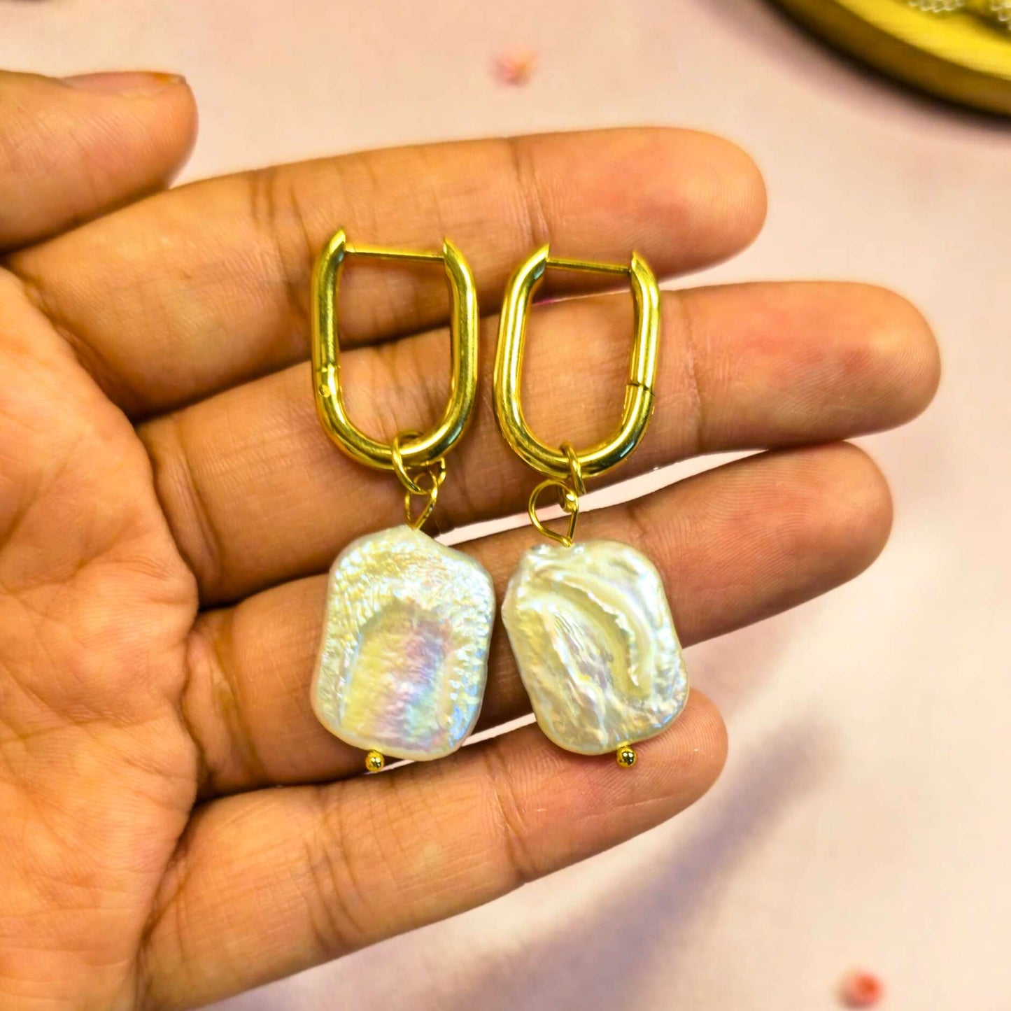 Gold hoop earrings with pearl held in a hand against a neutral background