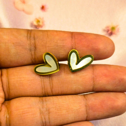 Gold heart-shaped white enamel centered earrings held between fingers with a blurred floral background