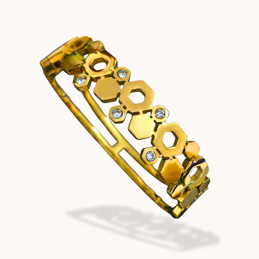 A gold-toned bangle bracelet featuring a repeating honeycomb pattern with small clear rhinestones, shown on a light off-white background with a soft shadow.