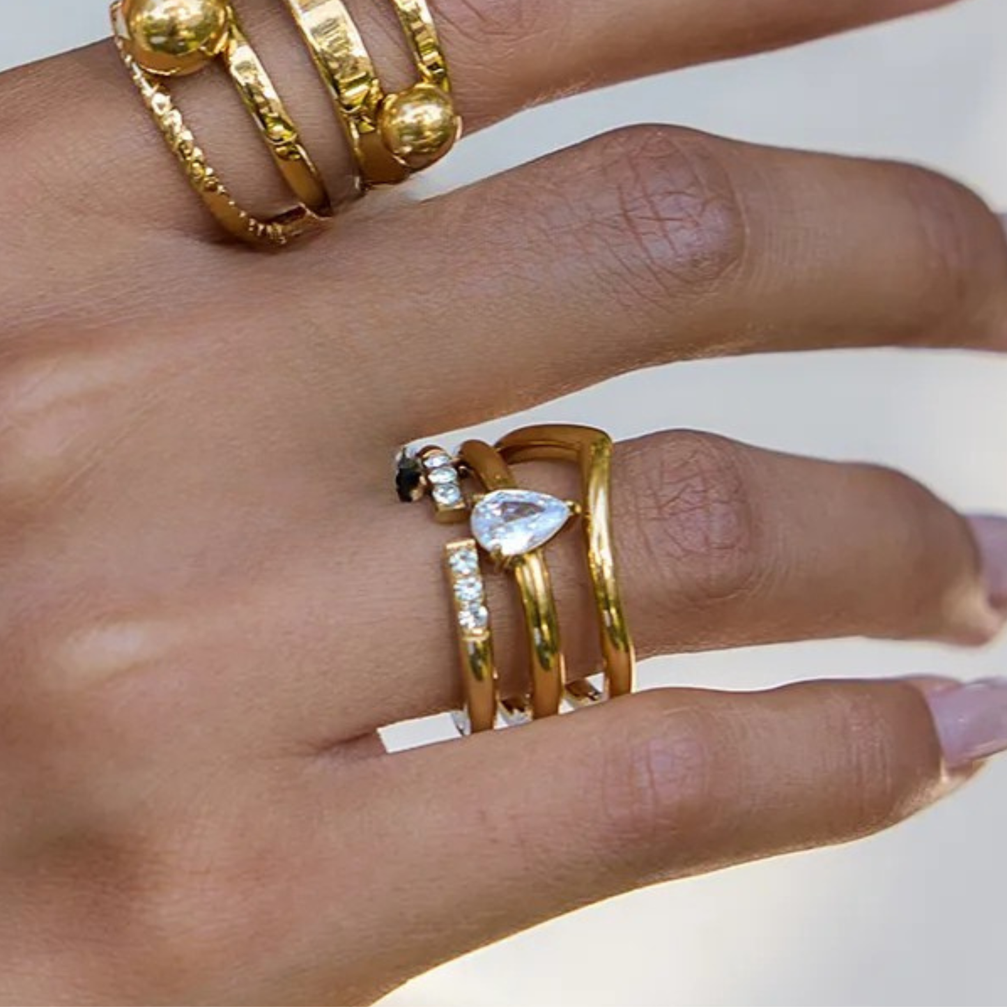 Close-up of a hand wearing multiple gold rings with clear gemstones on a neutral background