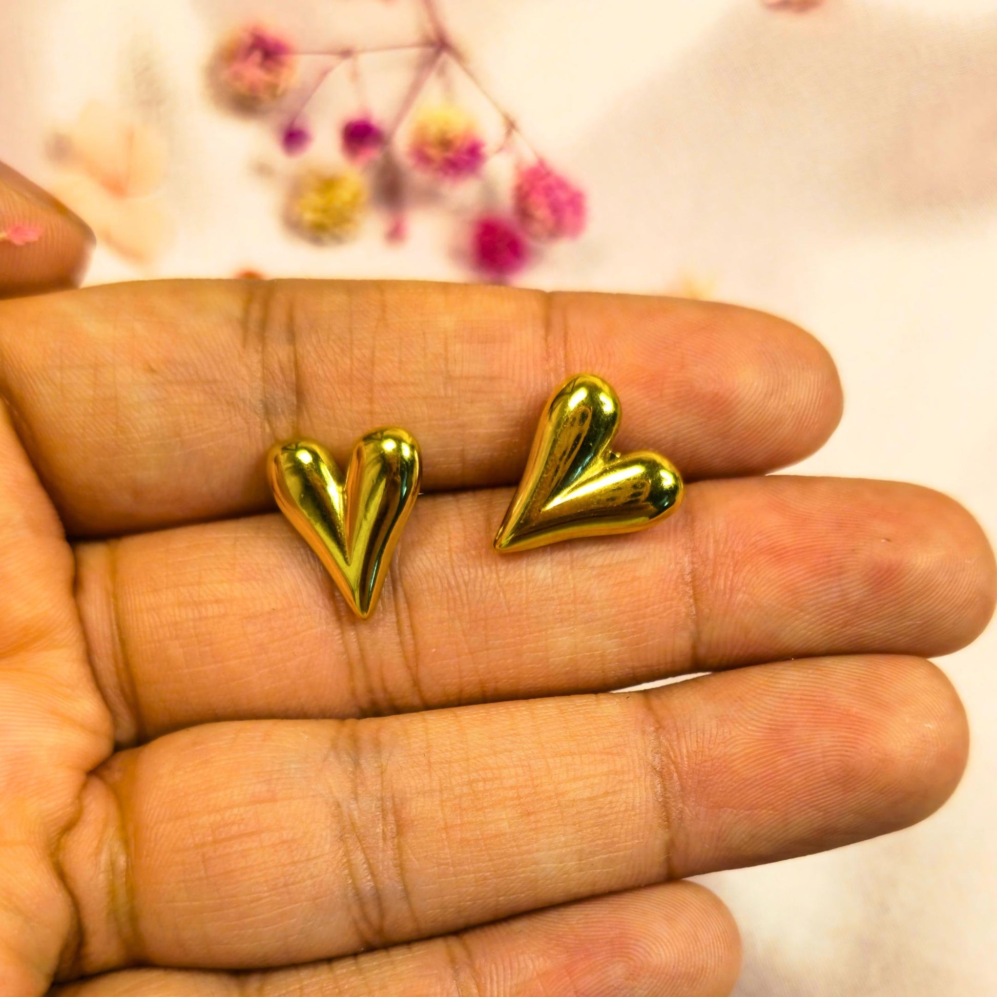 Gold heart-shaped earrings held between fingers with a blurred floral background