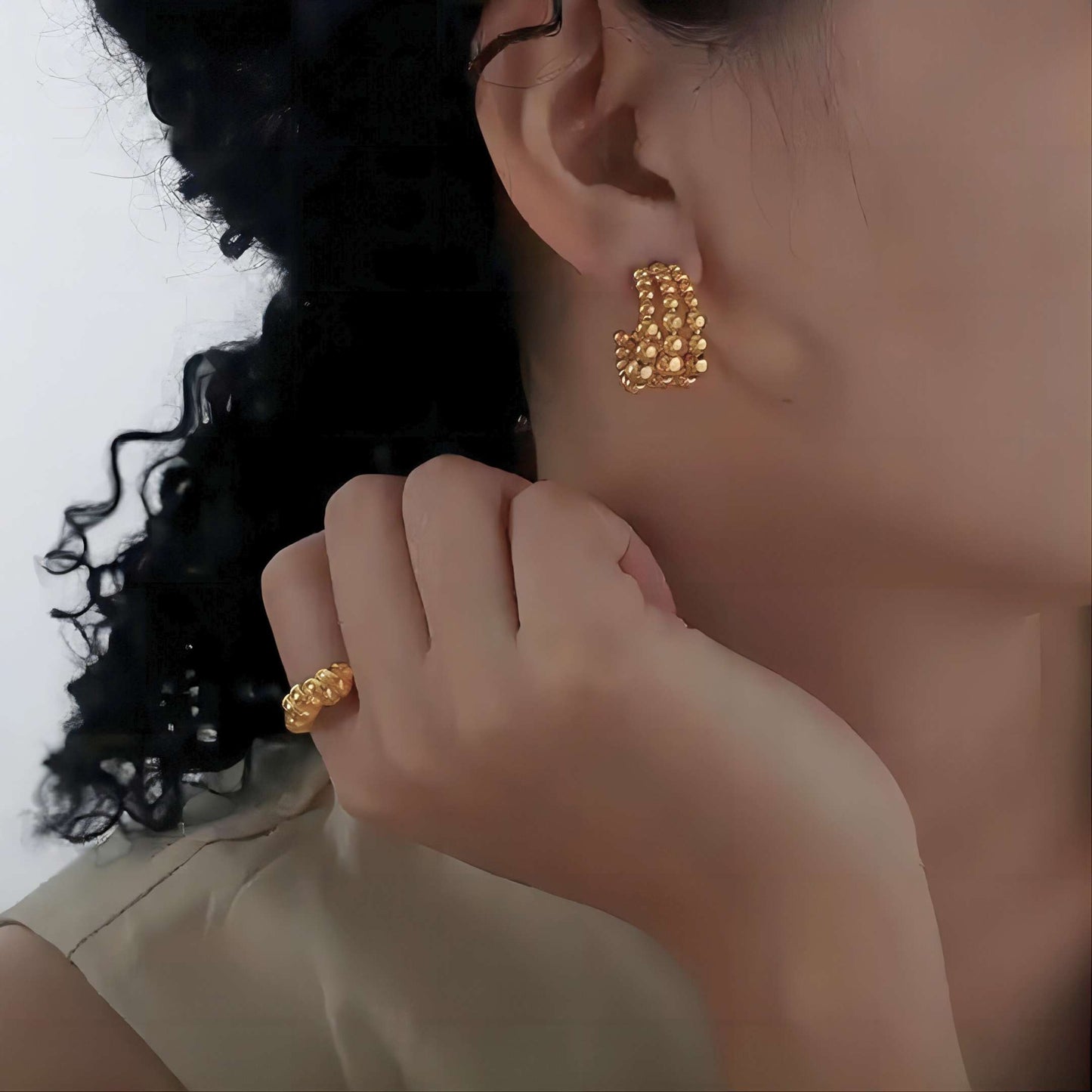 Close-up of a person wearing gold beaded earrings and a ring, with a neutral background.