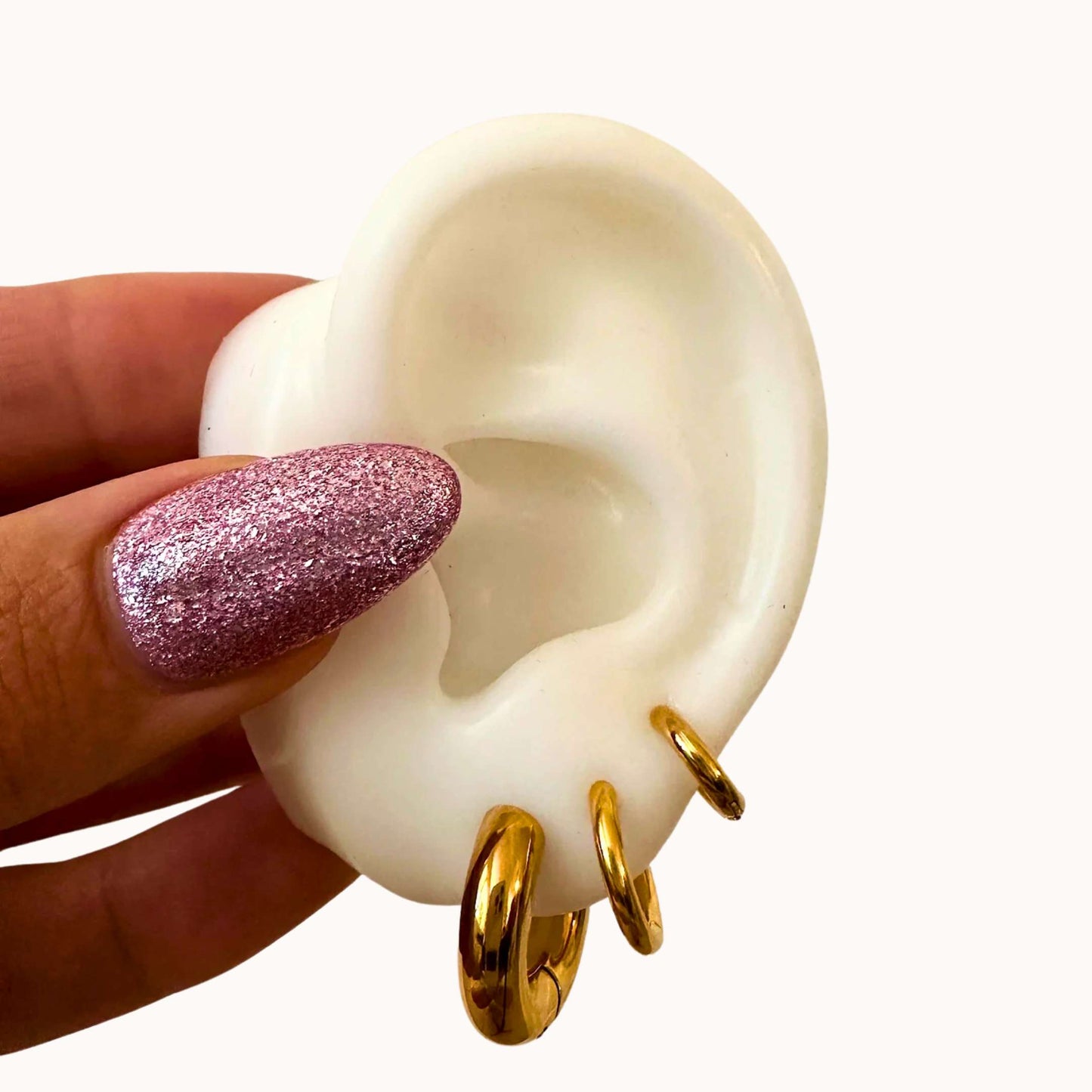 White ear model with gold earrings held by a hand with pink glittery nail polish.