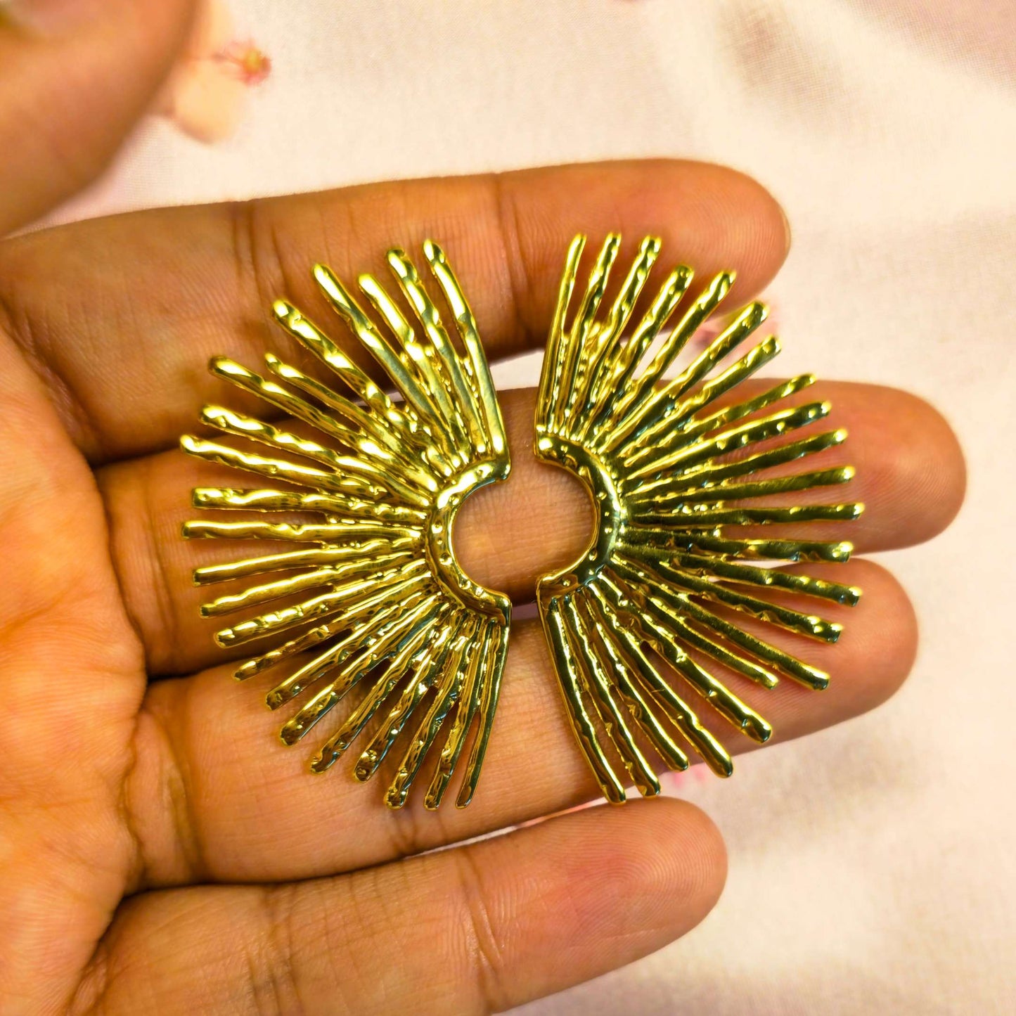 Gold sunburst design earring held between fingers against a light background 