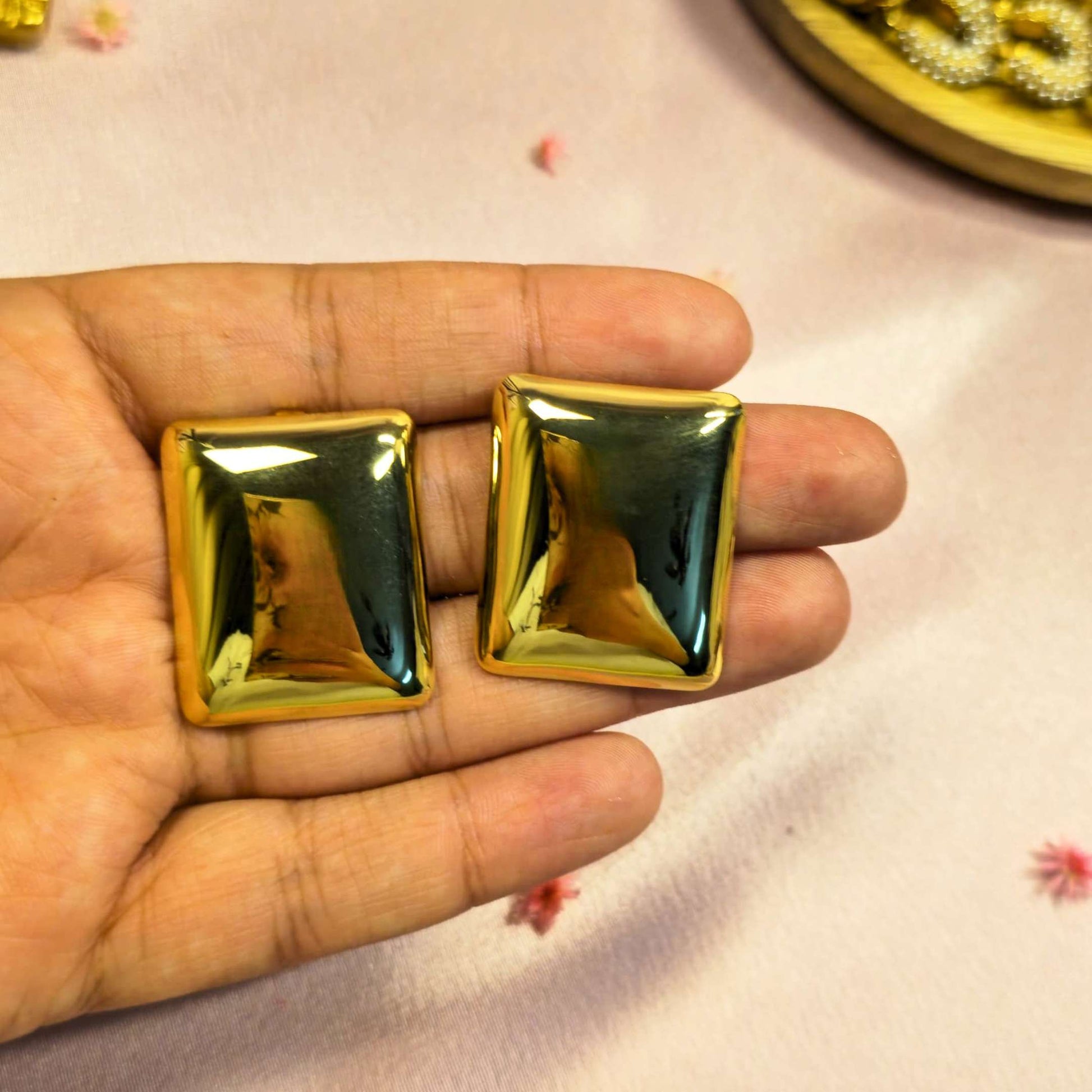 Gold square-shaped stud earring held in a hand on a light pink background