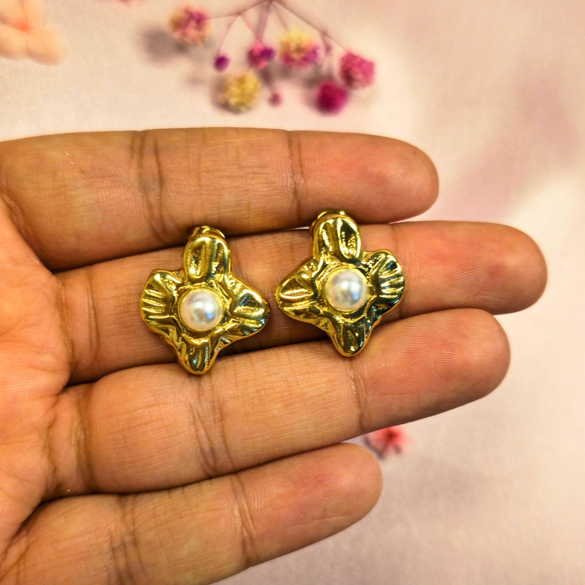 Gold earrings with pearl centers held between fingers against a blurred floral background