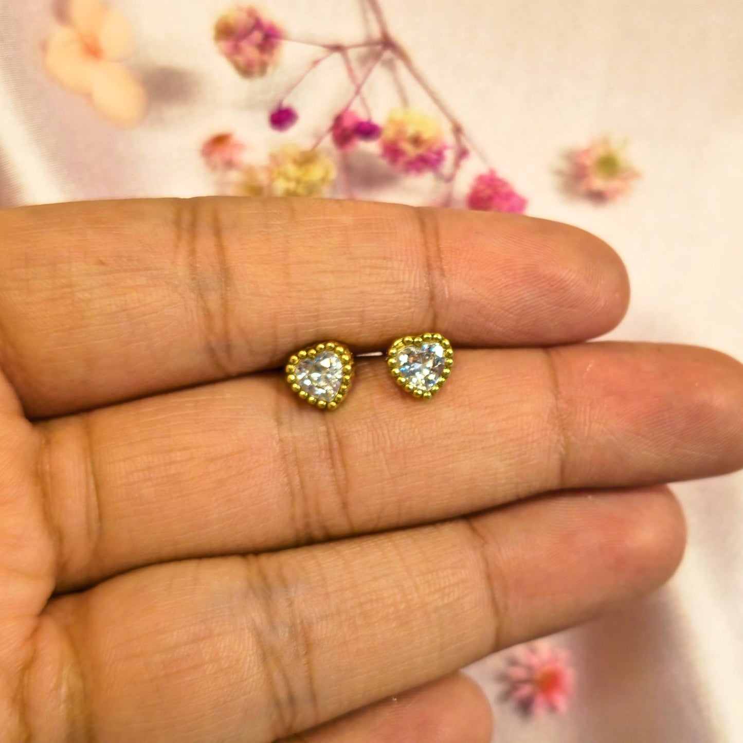 Gold Heart-shaped earrings held between fingers with a floral background
