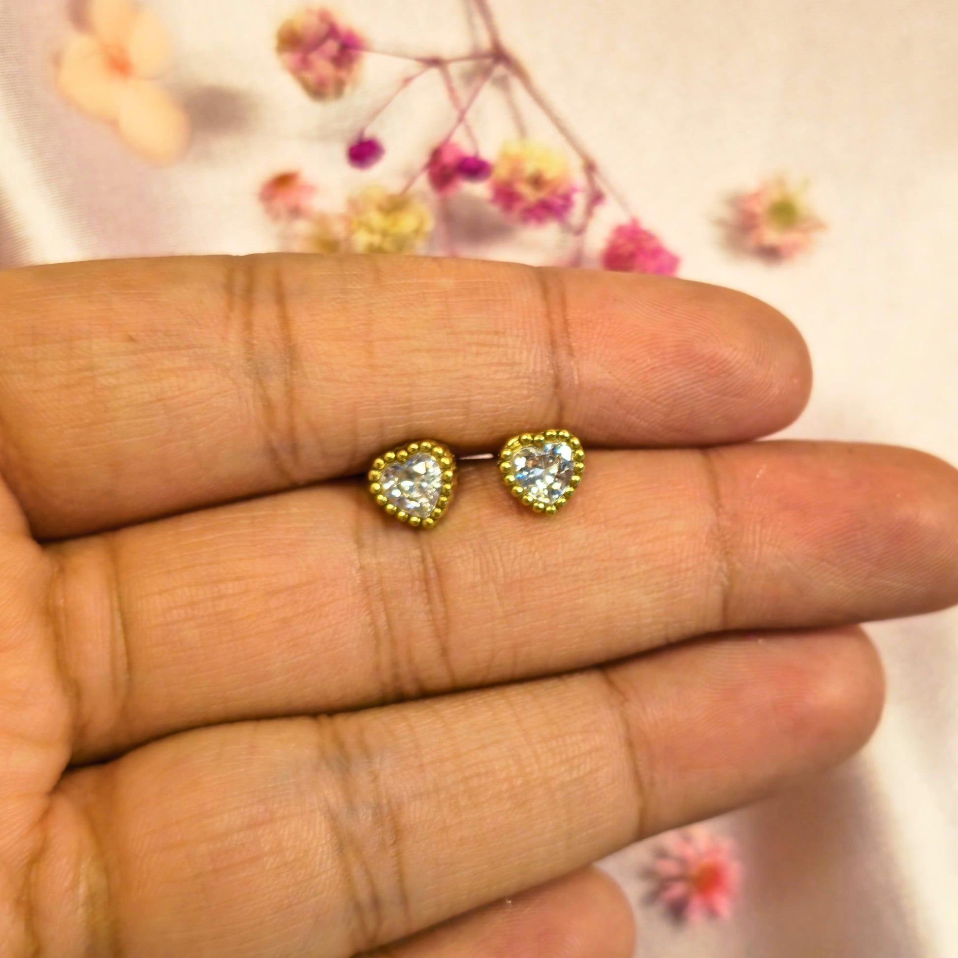 Gold Heart-shaped earrings held between fingers with a floral background