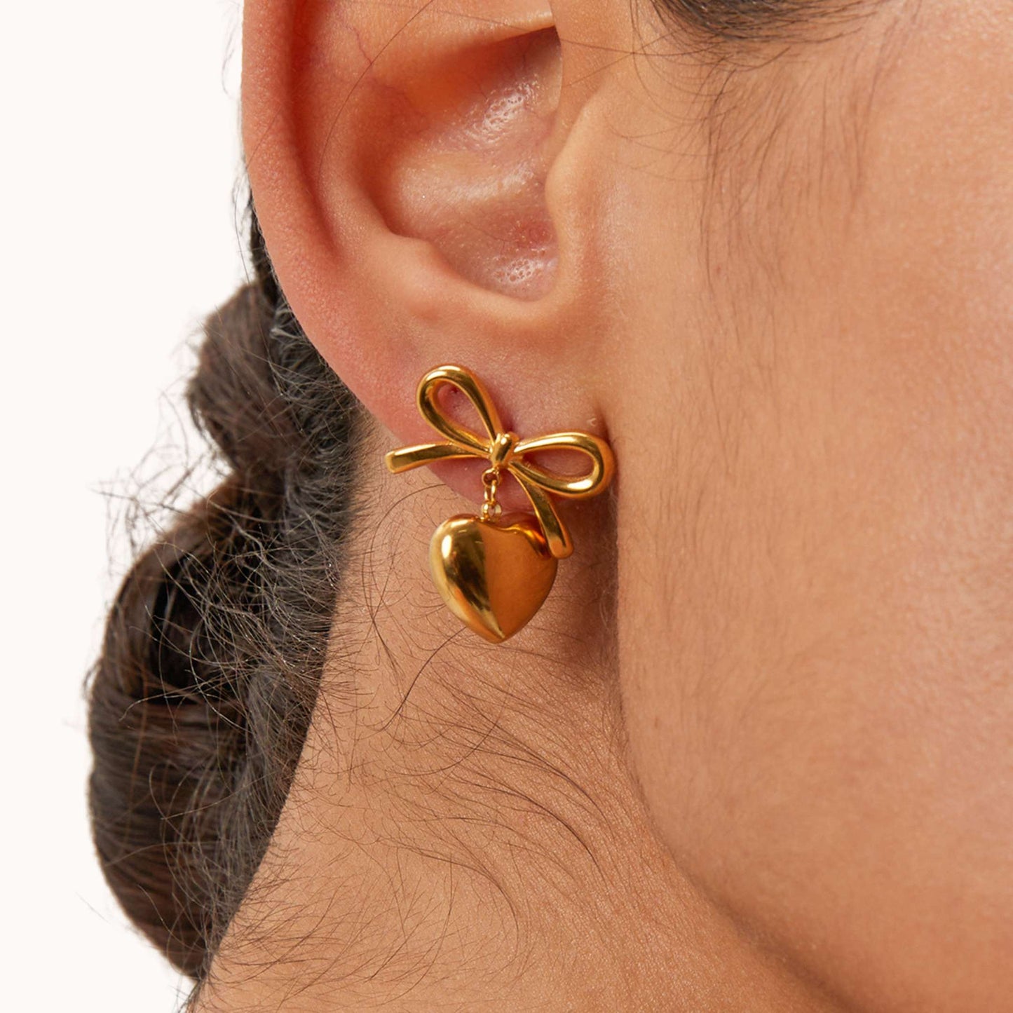 Gold Heart and bow shaped earring worn on an ear with a neutral background
