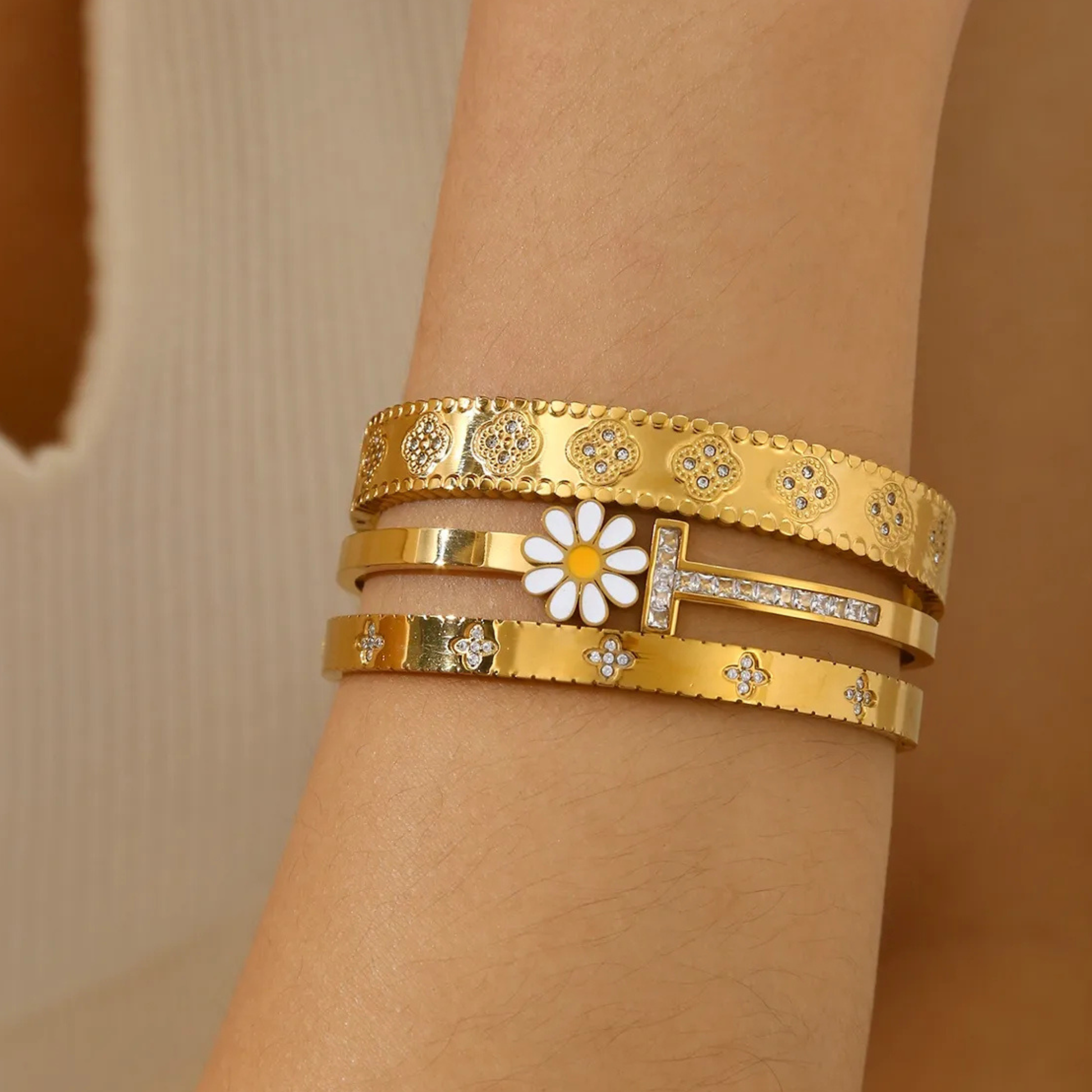 Gold bracelets with floral and letter 'T' design on a wrist against a beige background