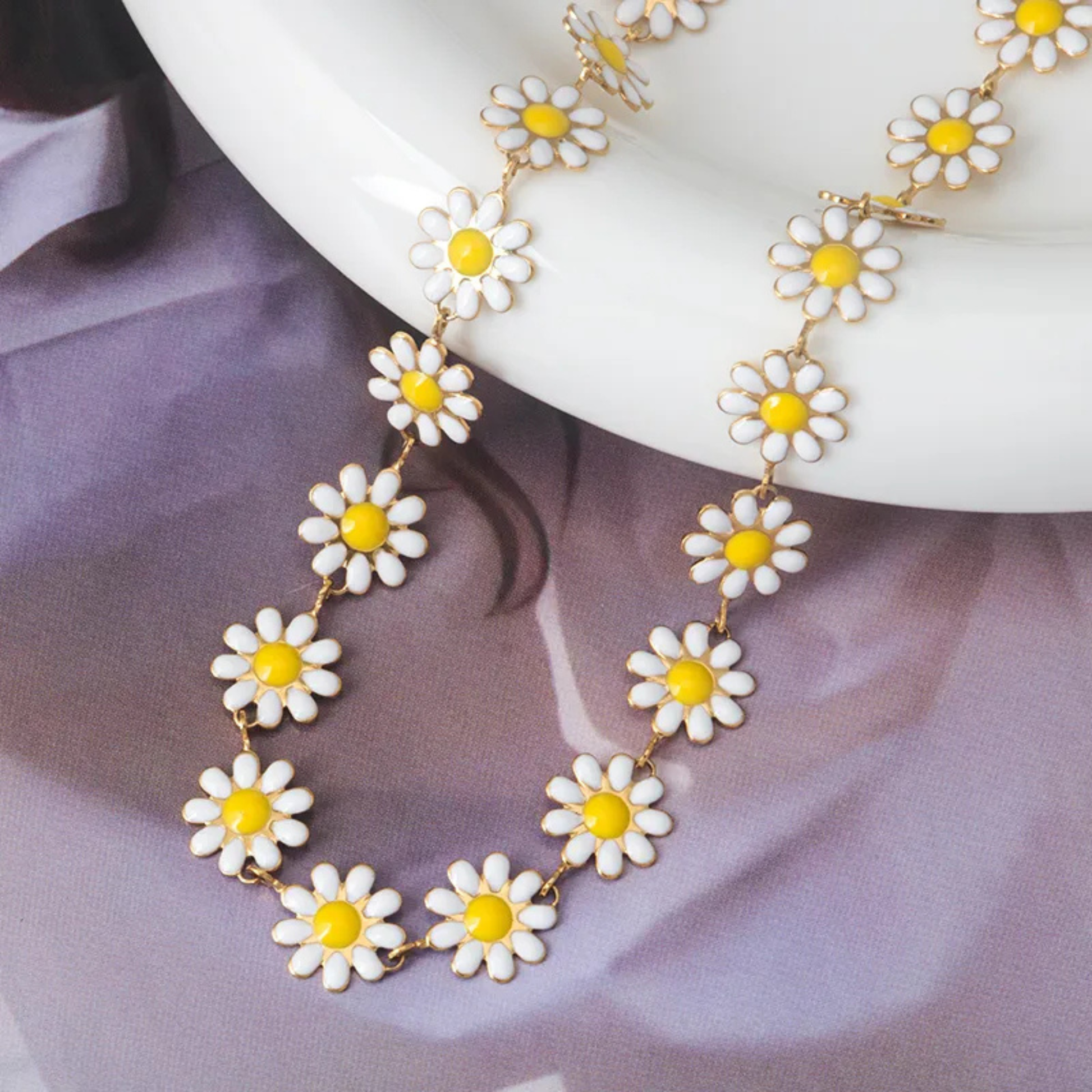 Daisy chain necklace with white flowers and yellow centers on a white plate against a purple fabric background.