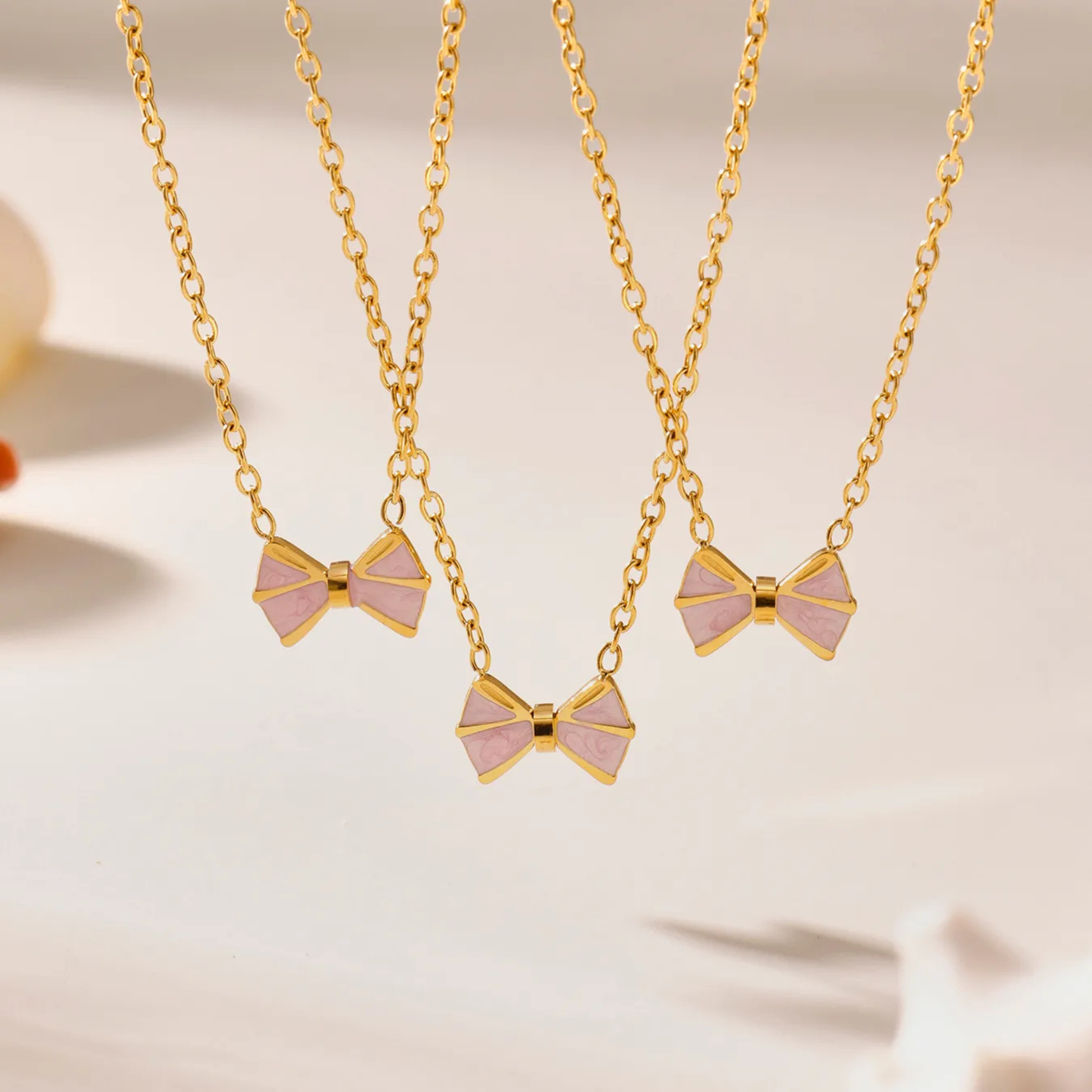 Two gold necklaces with pink bow pendants on a soft beige background