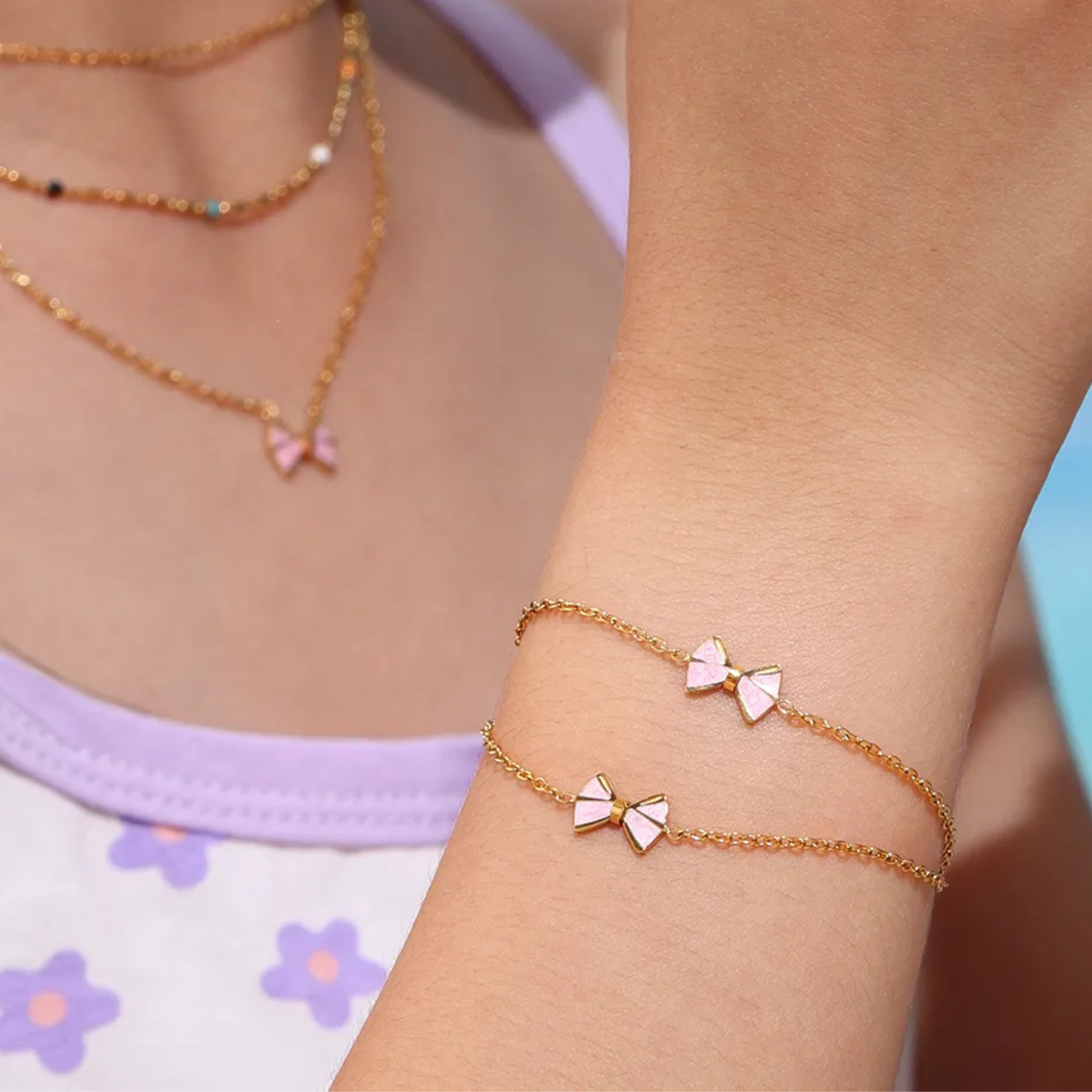 Gold bracelet with bow-shaped charms on a person's wrist, wearing a light purple top with floral patterns.