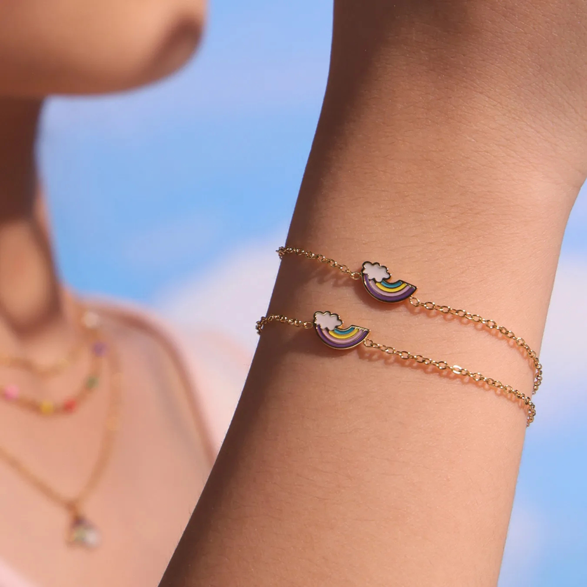 Person wearing a gold bracelet with cloud and rainbow design against a blue sky background