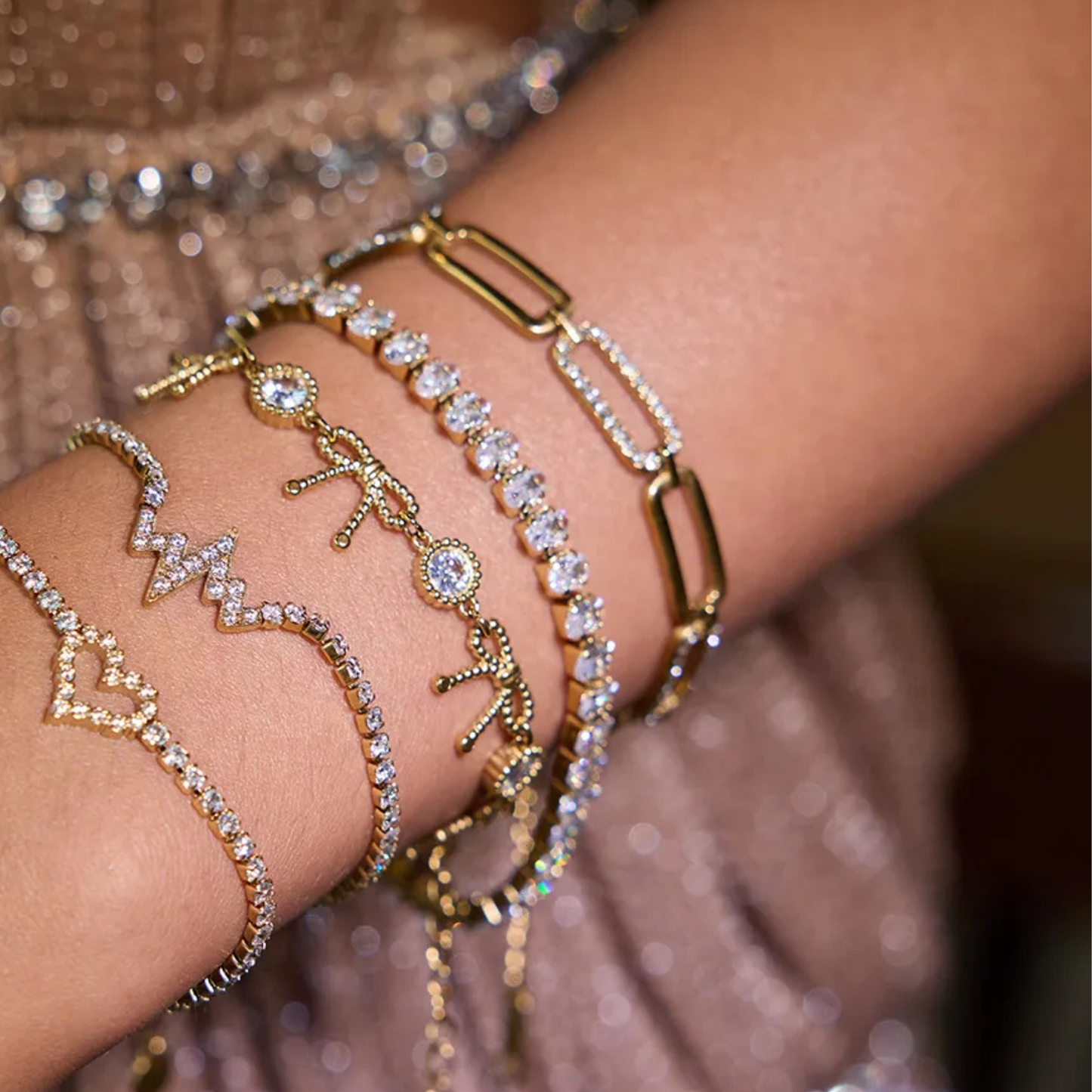 Close-up of a wrist wearing multiple gold and rose gold bracelets with various charms against a blurred background.