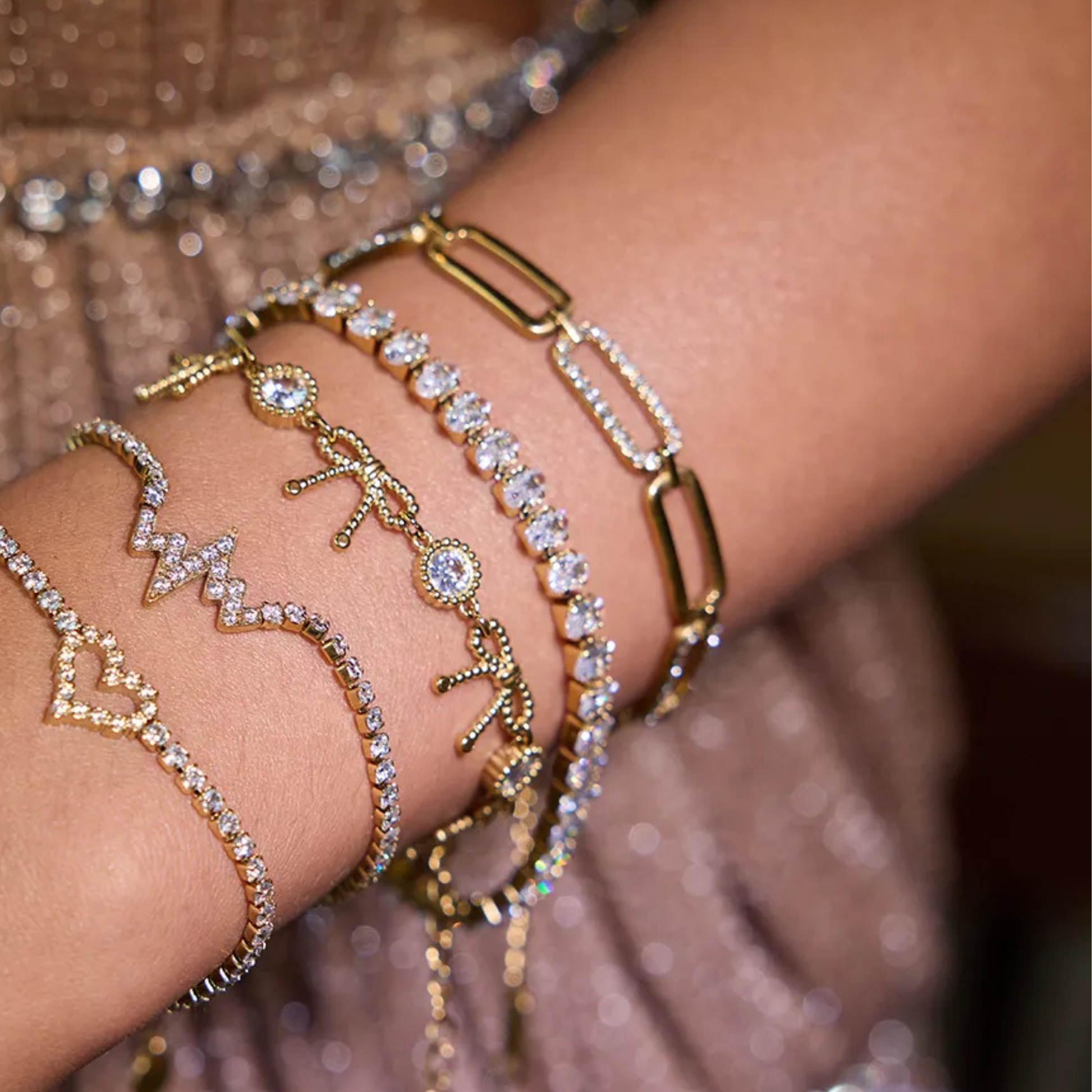 Close-up of a wrist wearing multiple gold and rose gold bracelets with various charms against a blurred background.