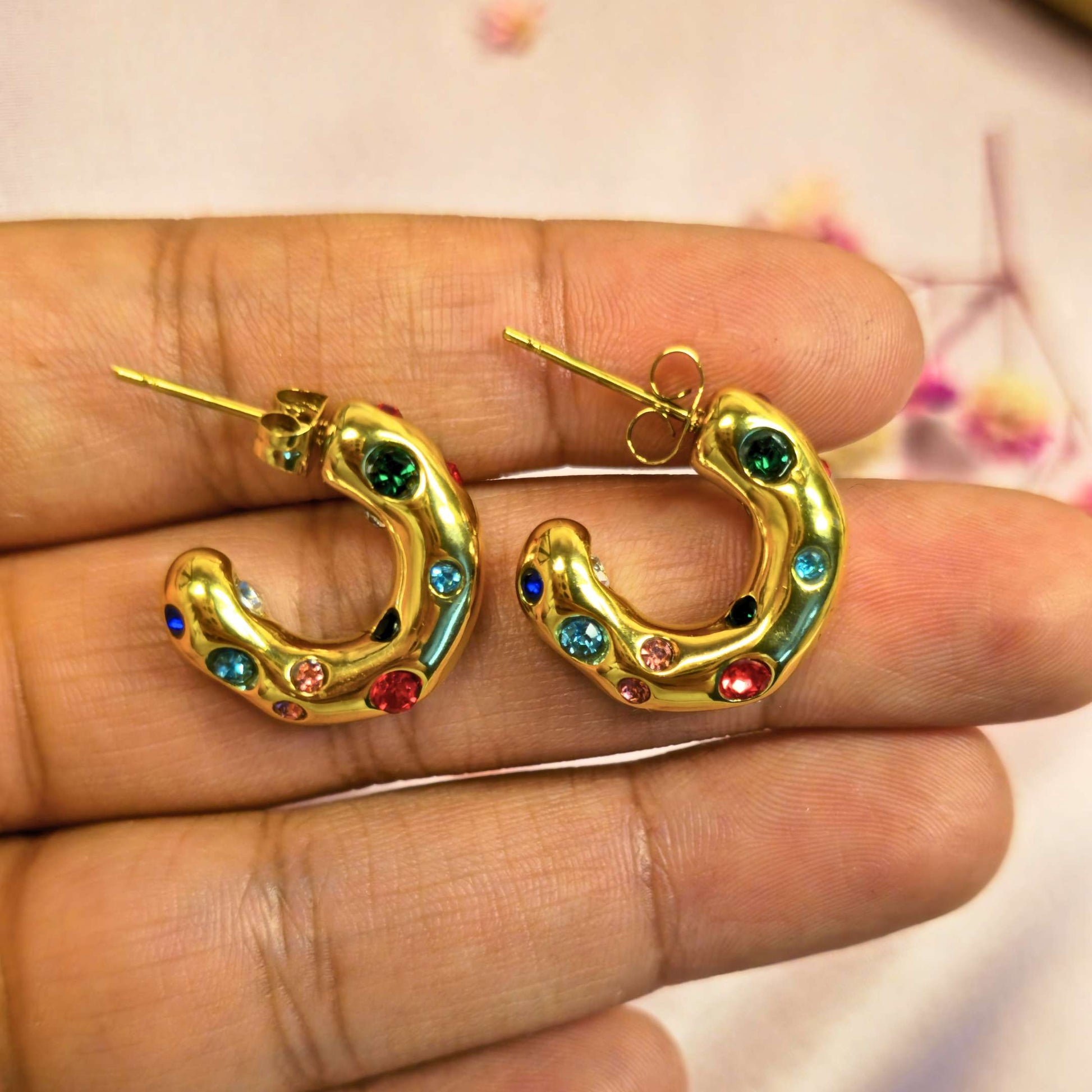 Gold hoop earrings with colorful gemstones held between fingers against a blurred background