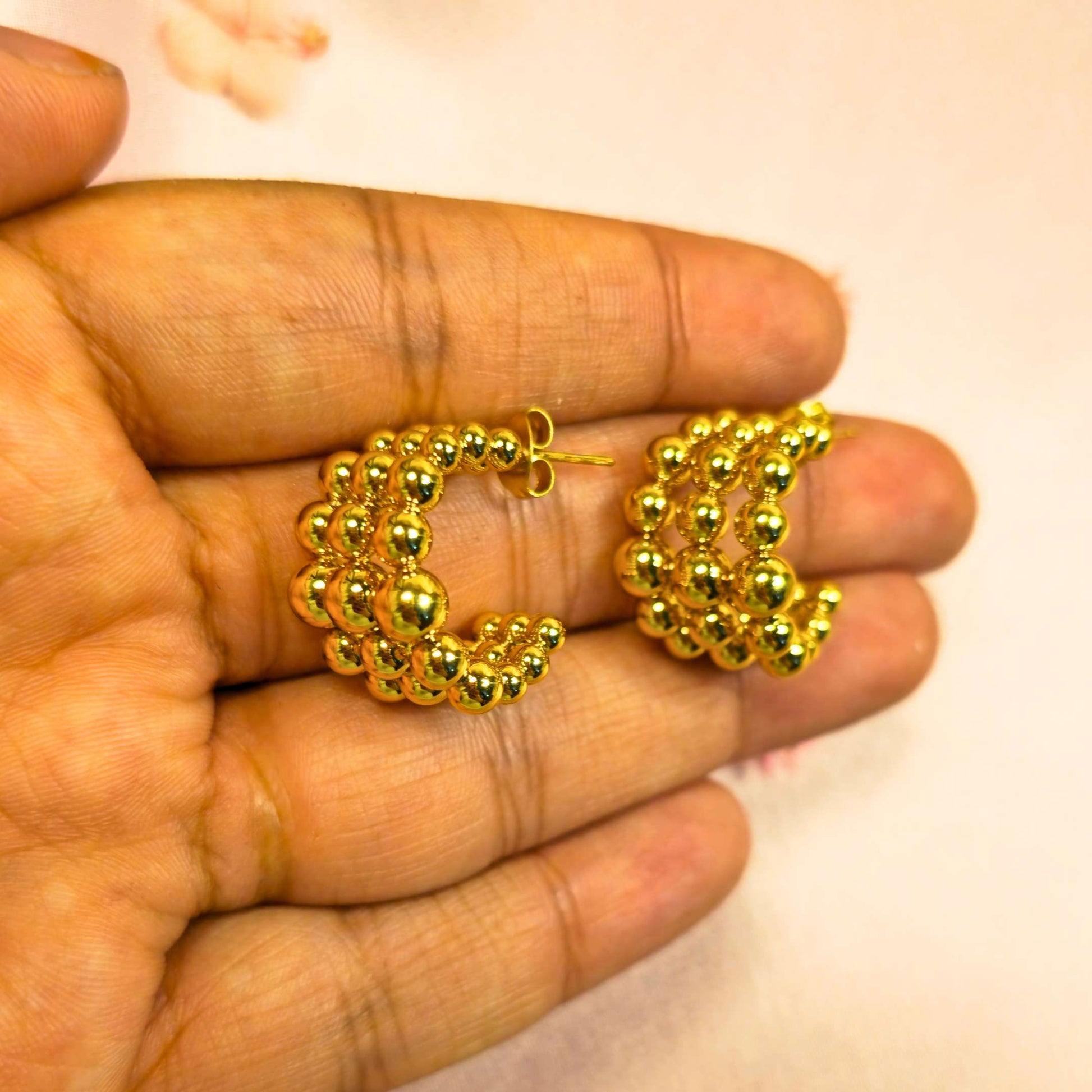 Gold beaded hoop earrings held between fingers against a neutral background