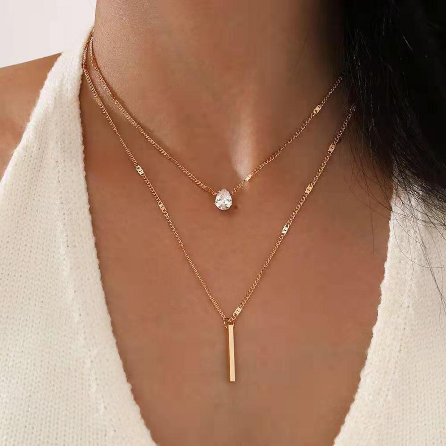 Two gold necklaces with a small round pendant and a bar pendant on a person wearing a white top.