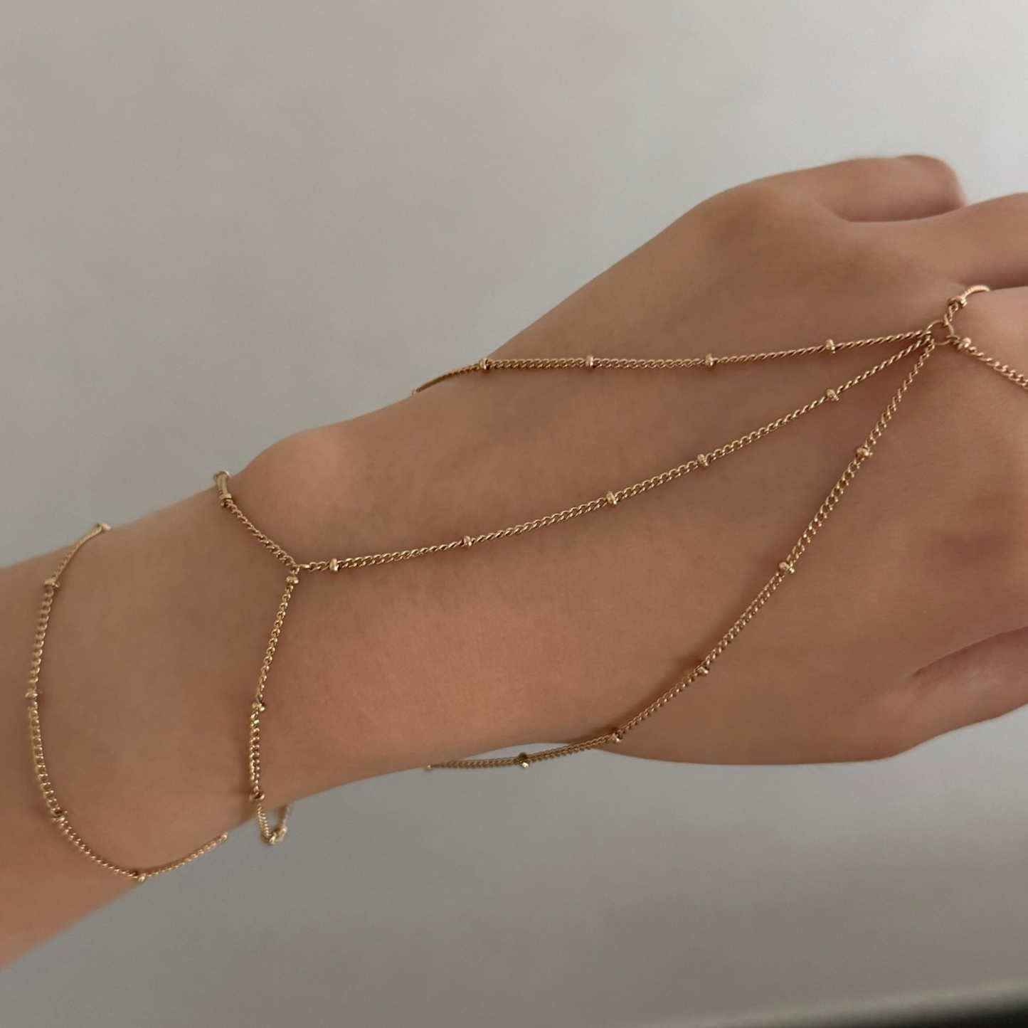 Hand wearing multiple gold ring chain bracelets on a neutral background