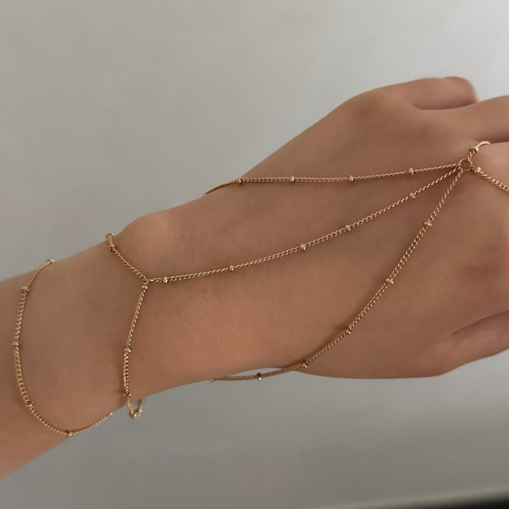 Hand wearing multiple gold ring chain bracelets on a neutral background