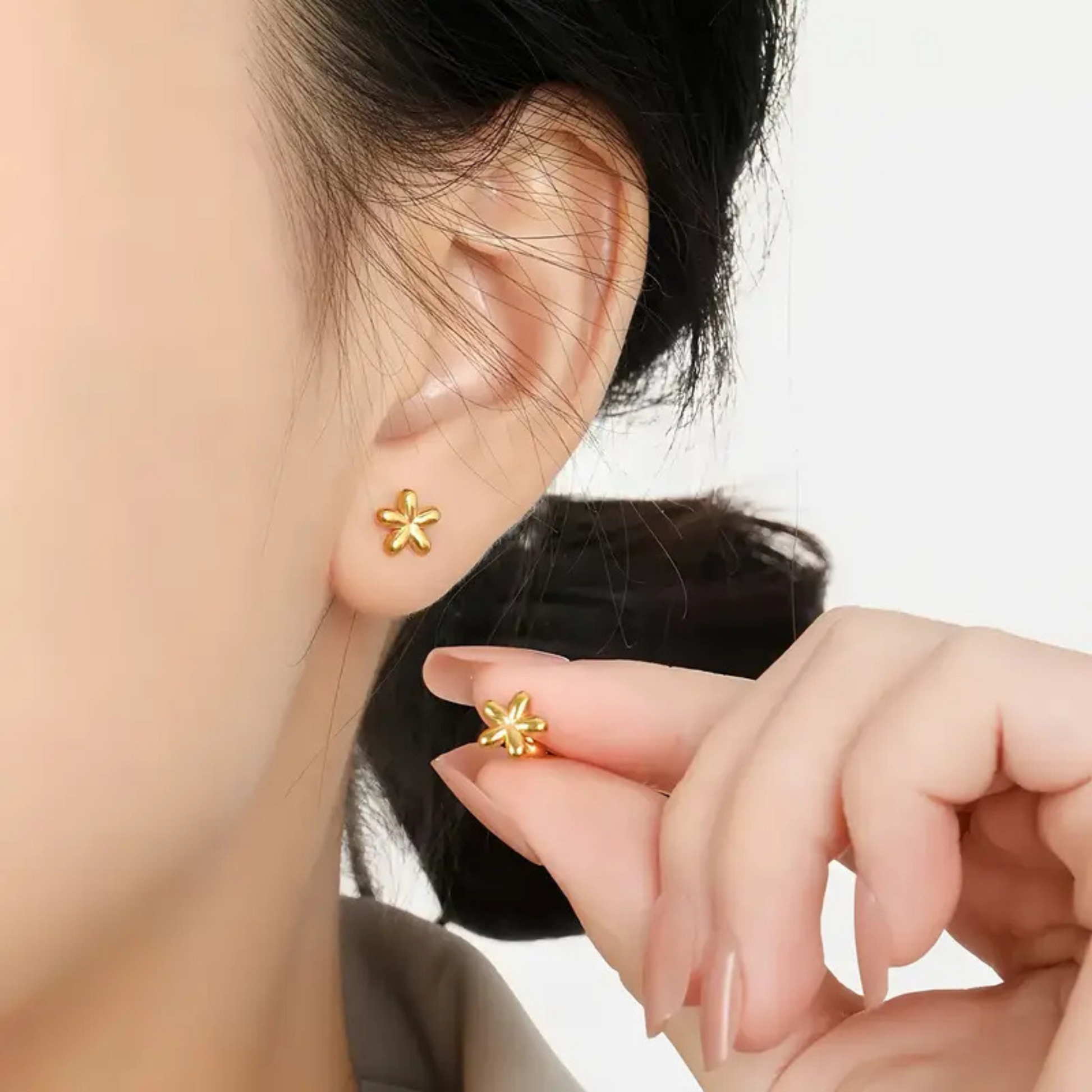 Gold flower-shaped earrings being worn and held by a person against a neutral background