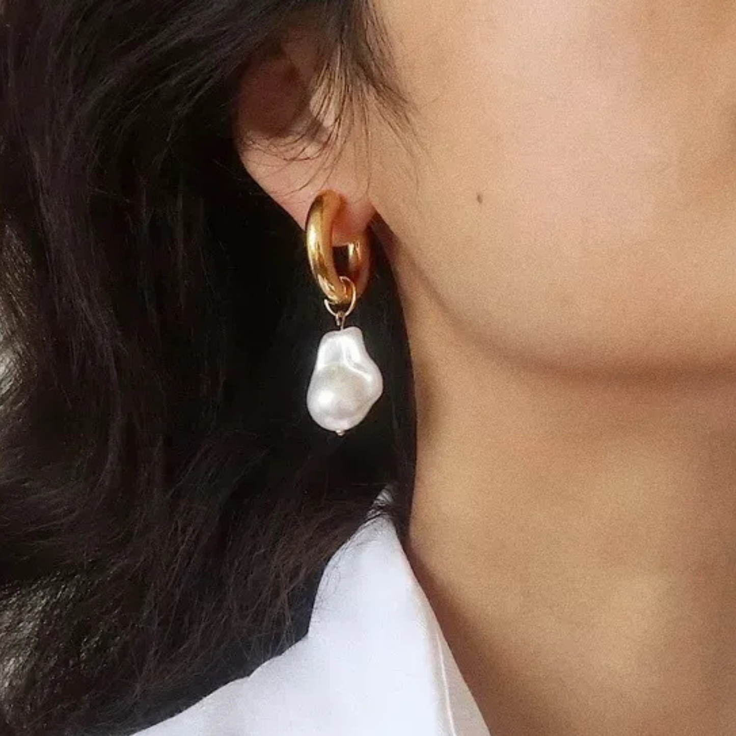 Close-up of a person wearing gold hoop earrings with white baroque pearl drops.