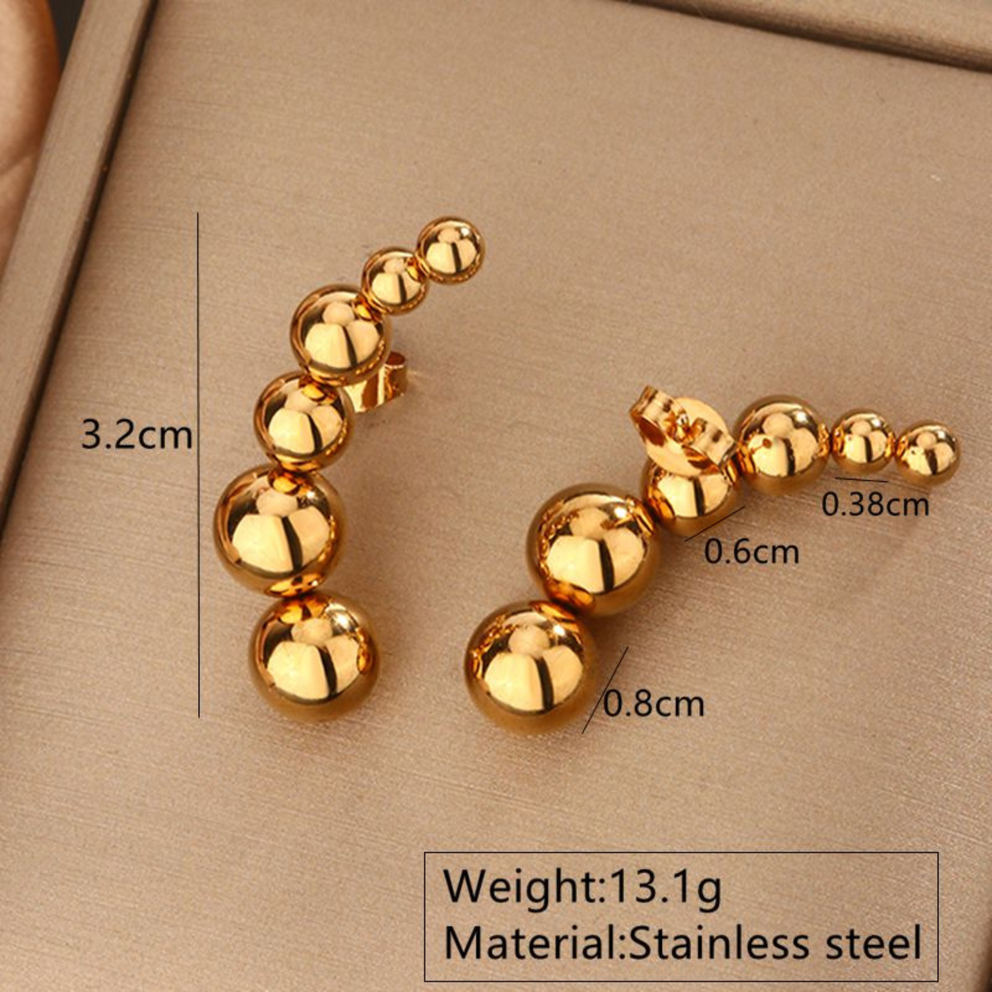Gold earrings with measurements and material details on a beige background