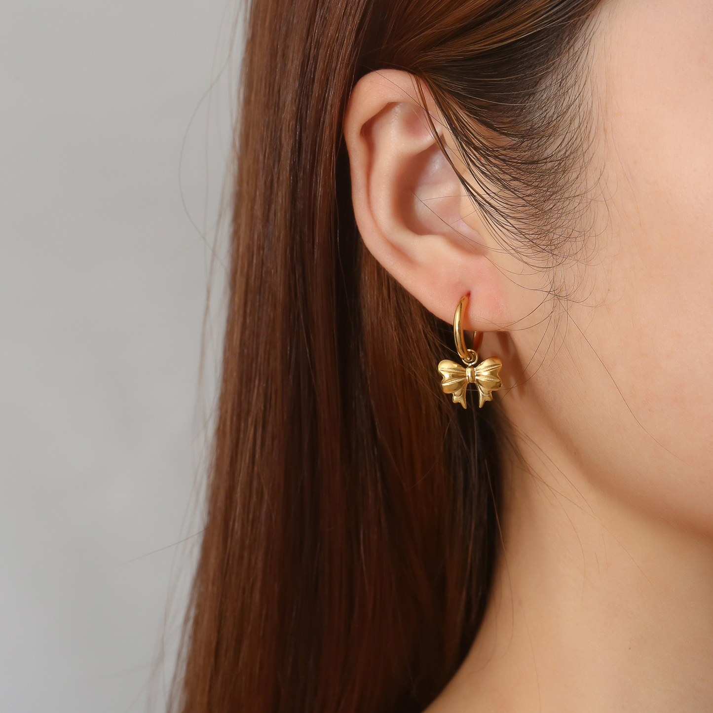 Gold bow-shaped earring worn by a person with long brown hair on a neutral background
