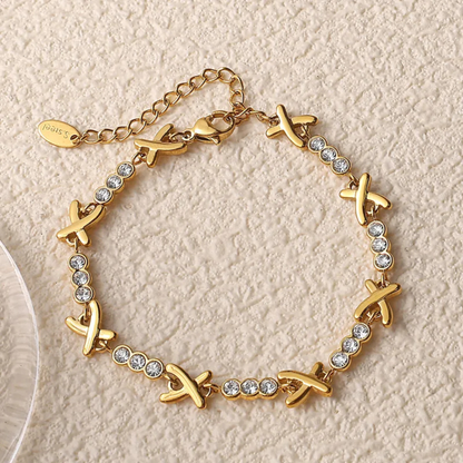 Gold bracelet with X-shaped charms and clear stones on a beige background
