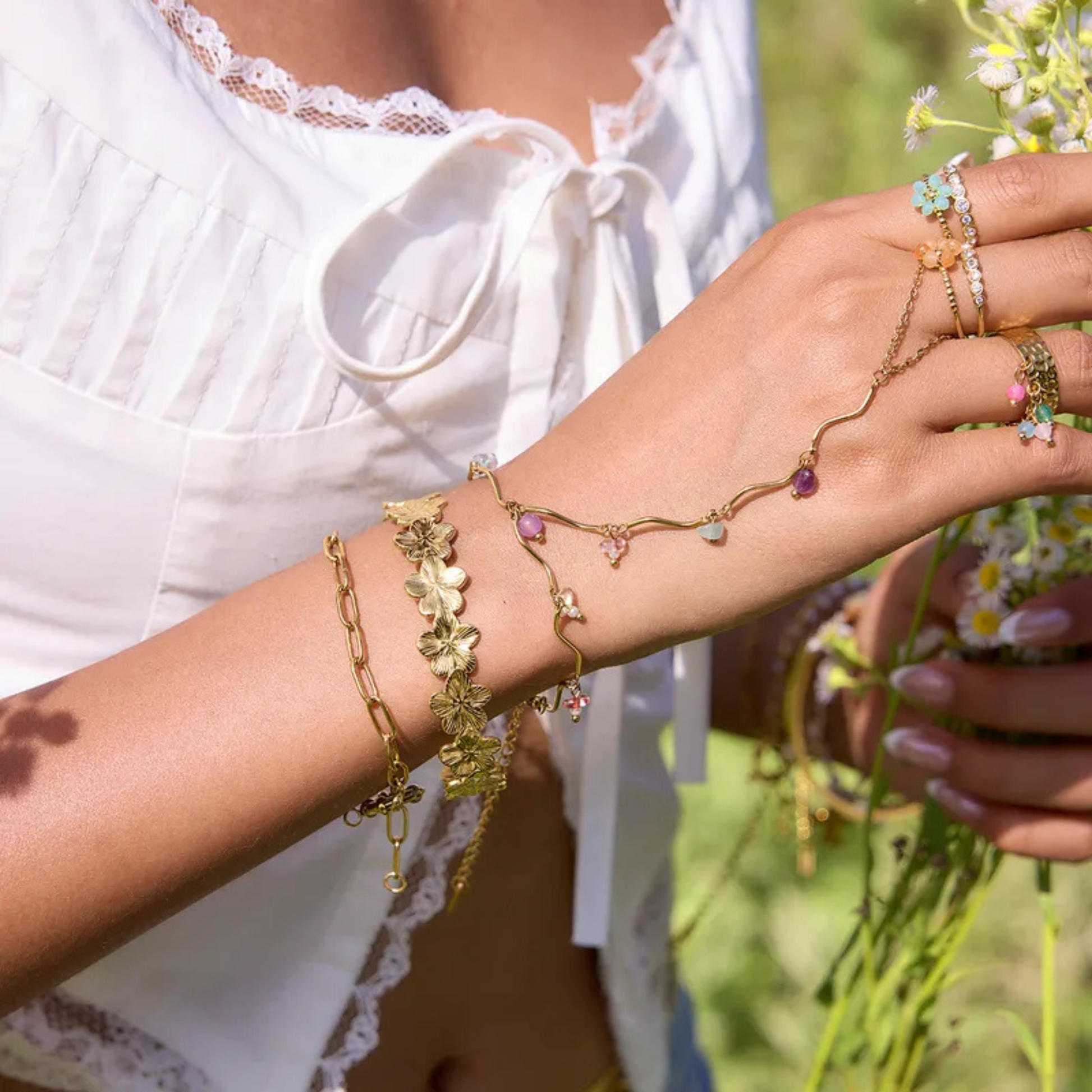 Close-up of a person's arm wearing multiple gold bracelets with gemstones, holding flowers.