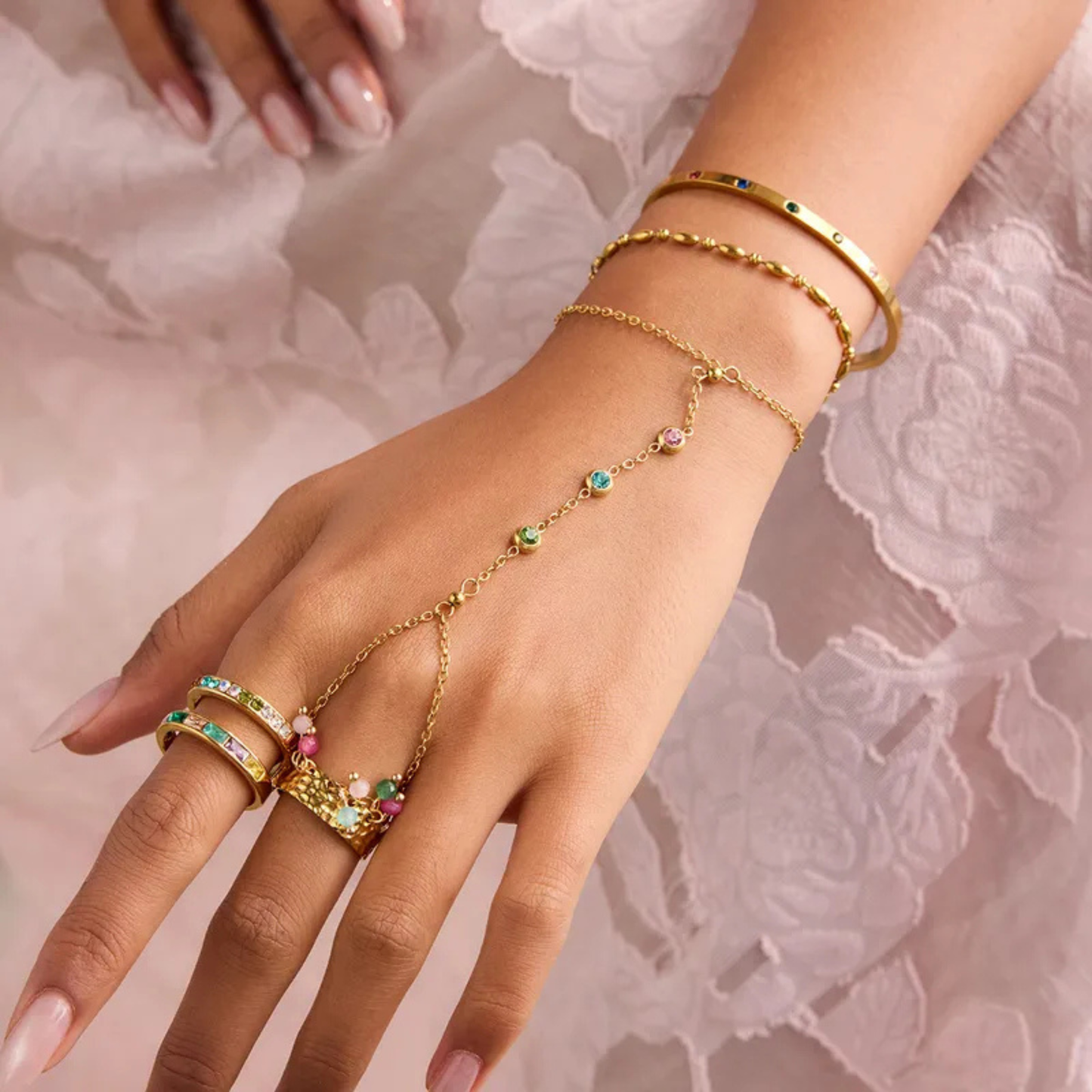 Gold jewelry set including rings and a bracelet on a hand against a soft pink lace background