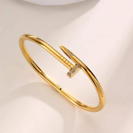 A polished gold hinged bangle bracelet with a barrette closure embellished with small clear stones, displayed on a soft cream-colored surface.