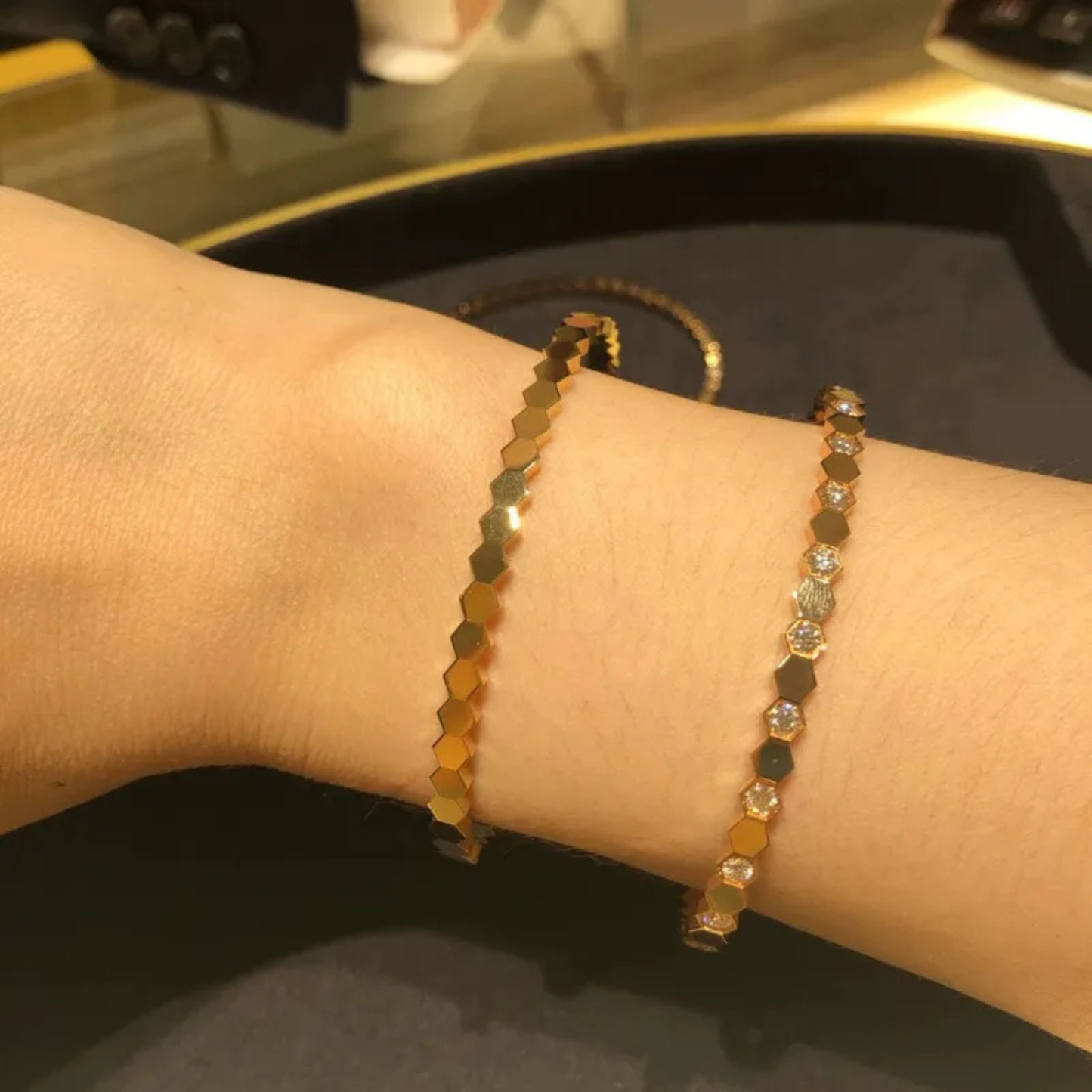 Close-up of a wrist wearing two gold bracelets on a reflective surface.