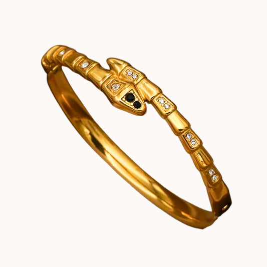 A gold snake bangle bracelet with small clear stones and a central black eye detail, shown on a plain off-white background.