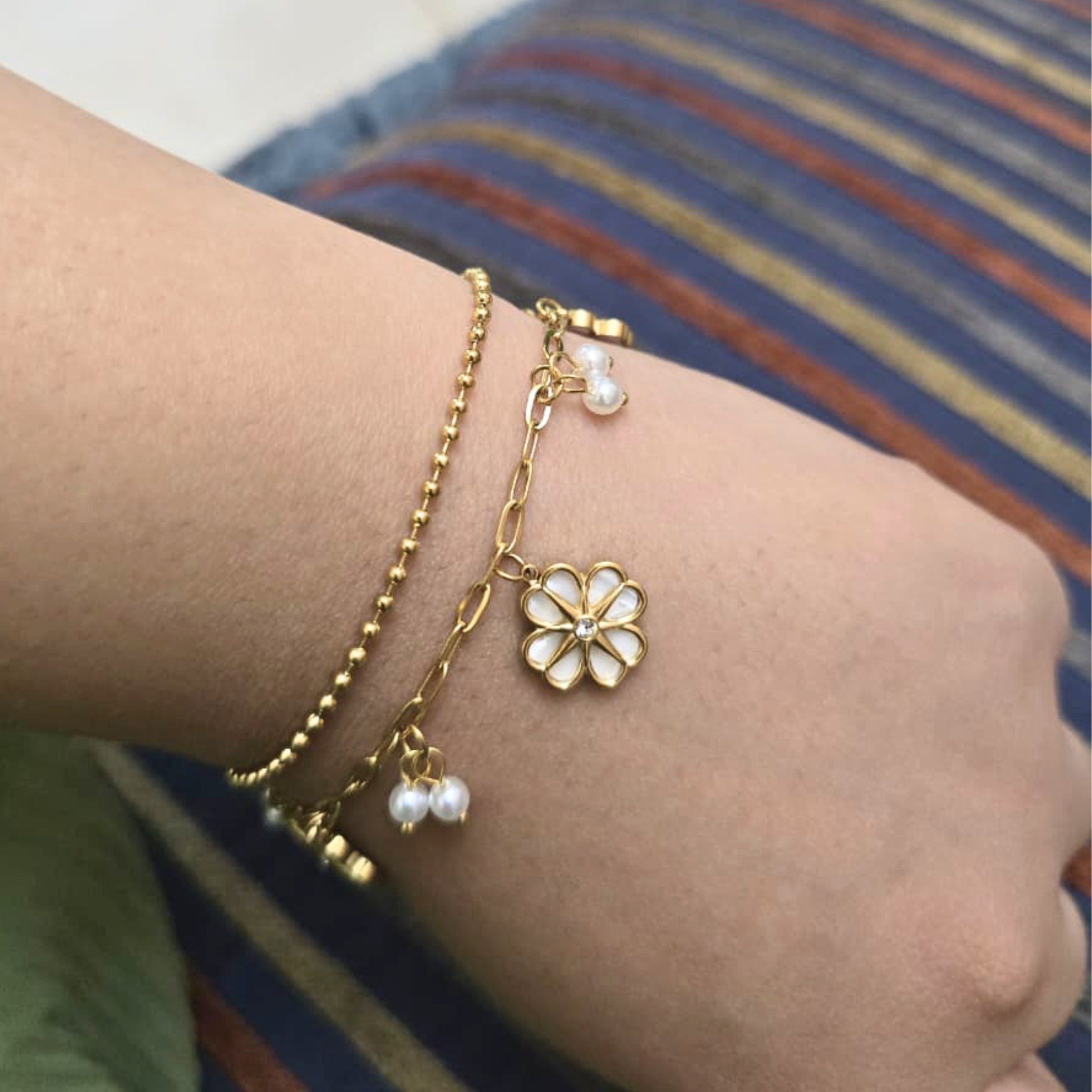 Gold bracelet with a flower charm and pearl beads on a wrist against a striped background