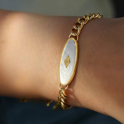Gold bracelet with a white oval charm on a wrist against a neutral background