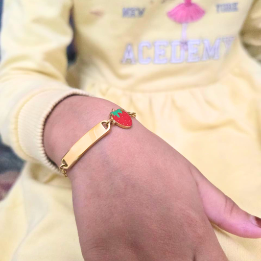 Gold bracelet with a red heart charm on a child's wrist, wearing a yellow outfit with text.